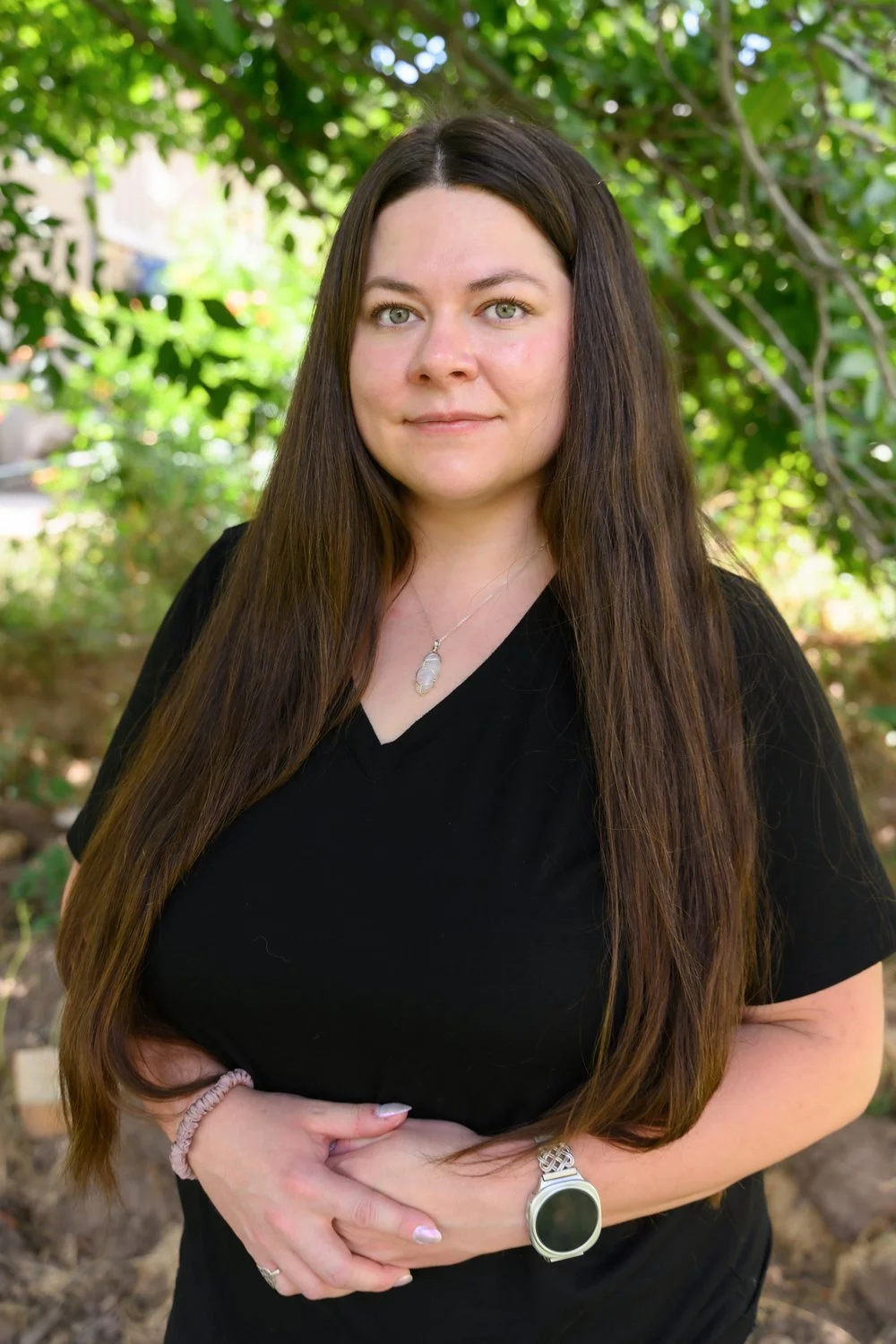 A woman with long brown hair and blue eyes, wearing a black top, standing outdoors in a green, leafy area with sunlight filtering through the trees.