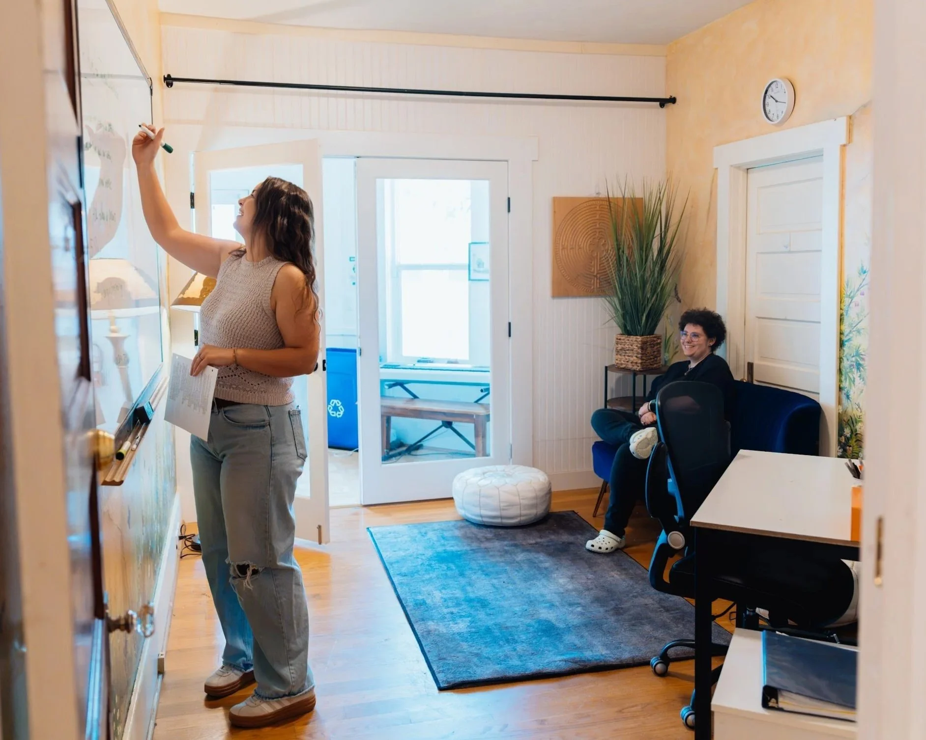 A woman writing on a whiteboard in a cozy, well-lit room with another woman sitting on a blue couch, smiling, and a large plant nearby.