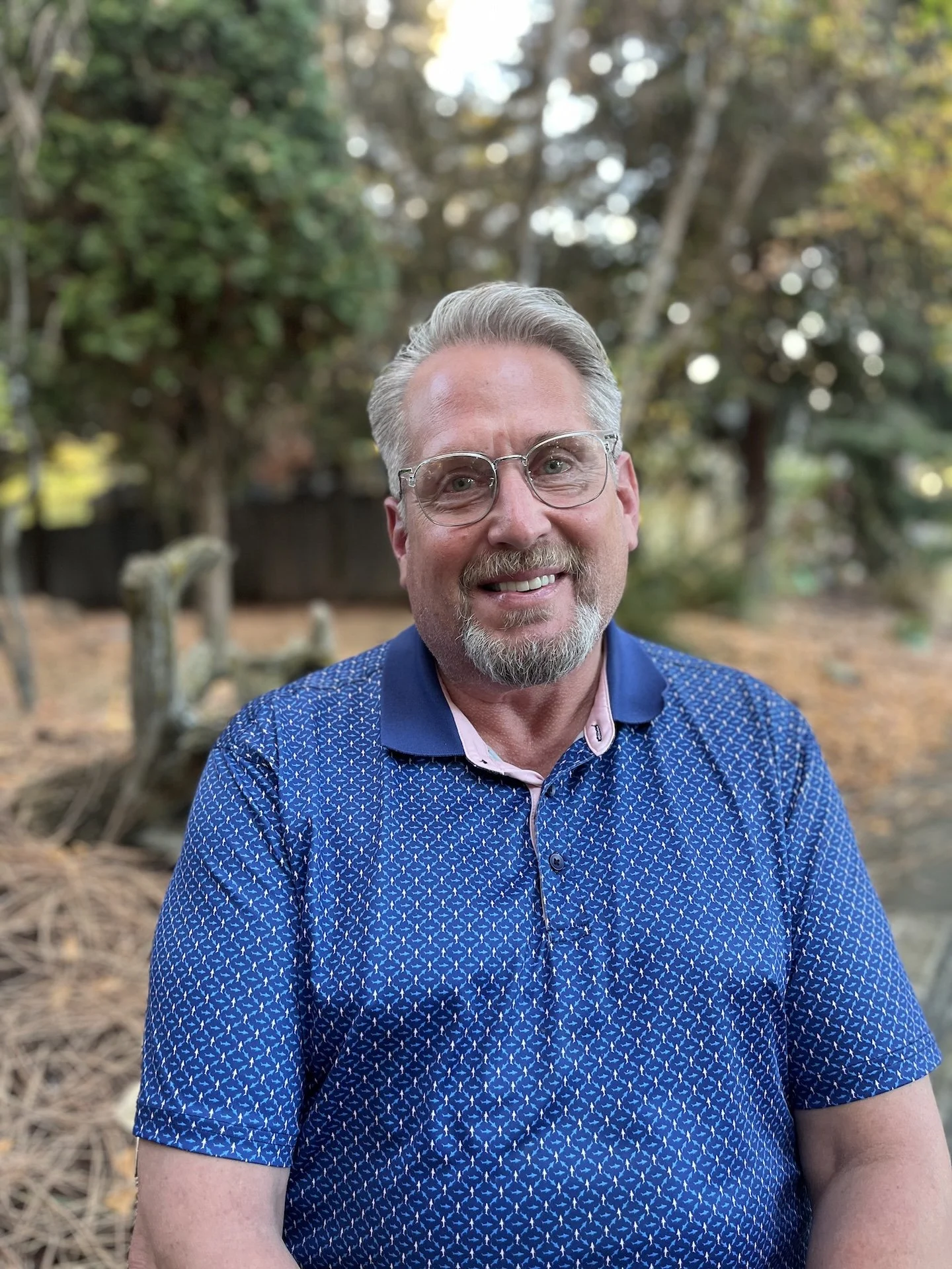 Portrait of a smiling middle-aged man with glasses, gray hair, and a beard, wearing a blue patterned shirt outdoors with trees and natural background.