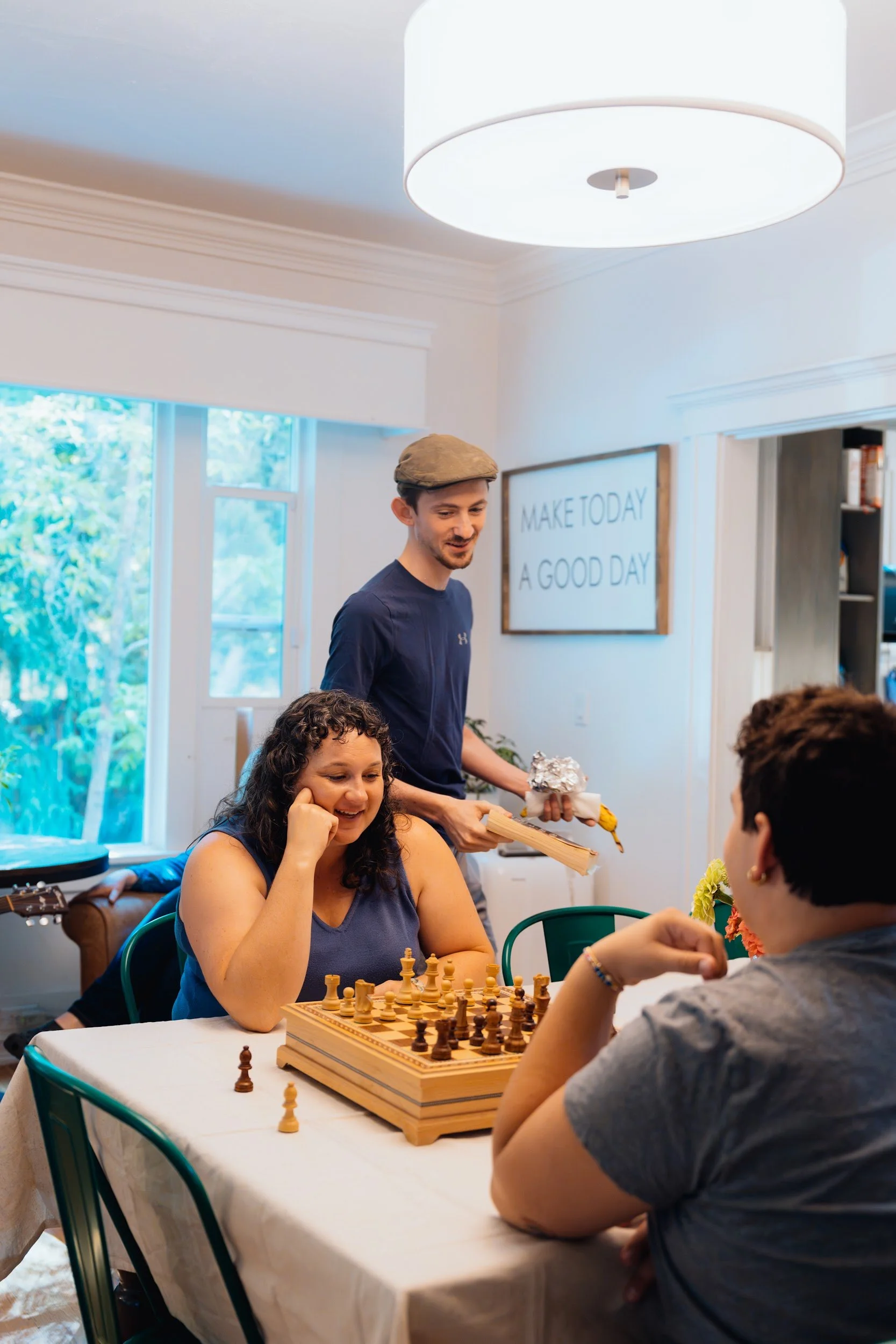 Young man in a flat cap smiling while friends sit at a kitchen table