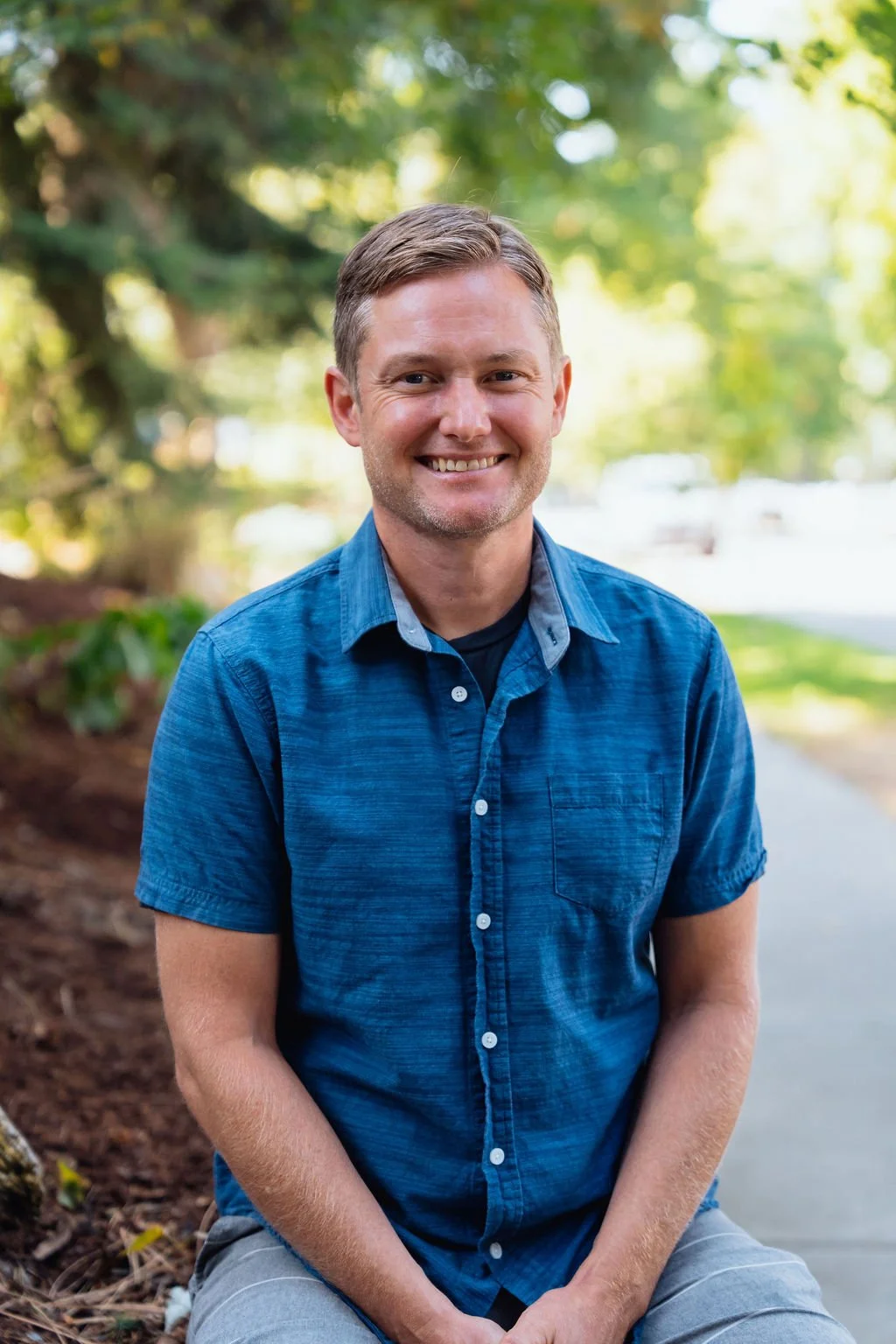 A smiling man in a blue short-sleeve button-up shirt sitting outdoors on a pathway, with trees and sunlight in the background.