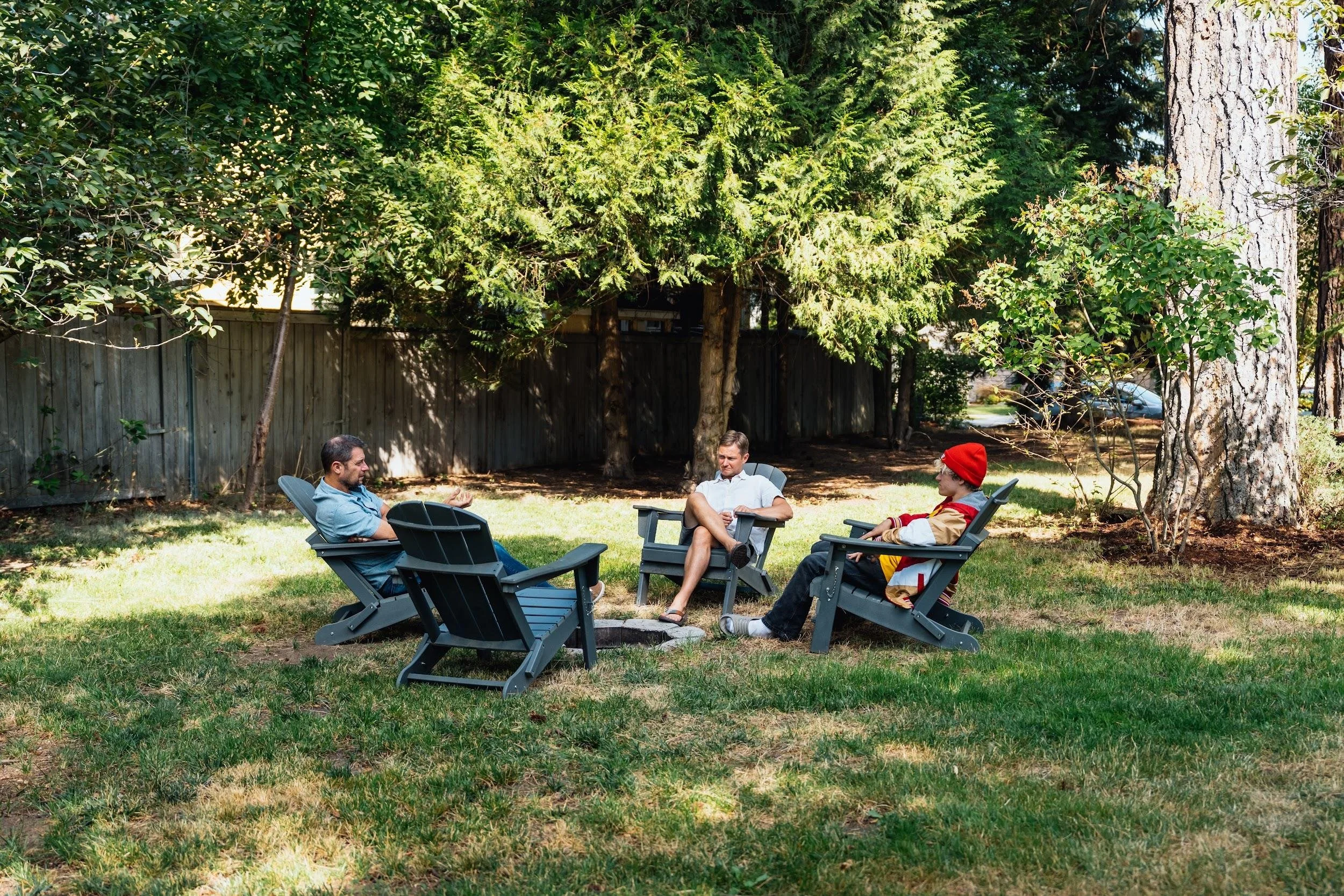 Four men sitting in outdoor chairs in a backyard, engaging in conversation, surrounded by trees and a wooden fence on a sunny day.