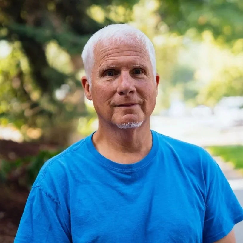A middle-aged man with short white hair and a goatee, wearing a bright blue T-shirt, standing outdoors with blurred green trees and sunlight in the background.