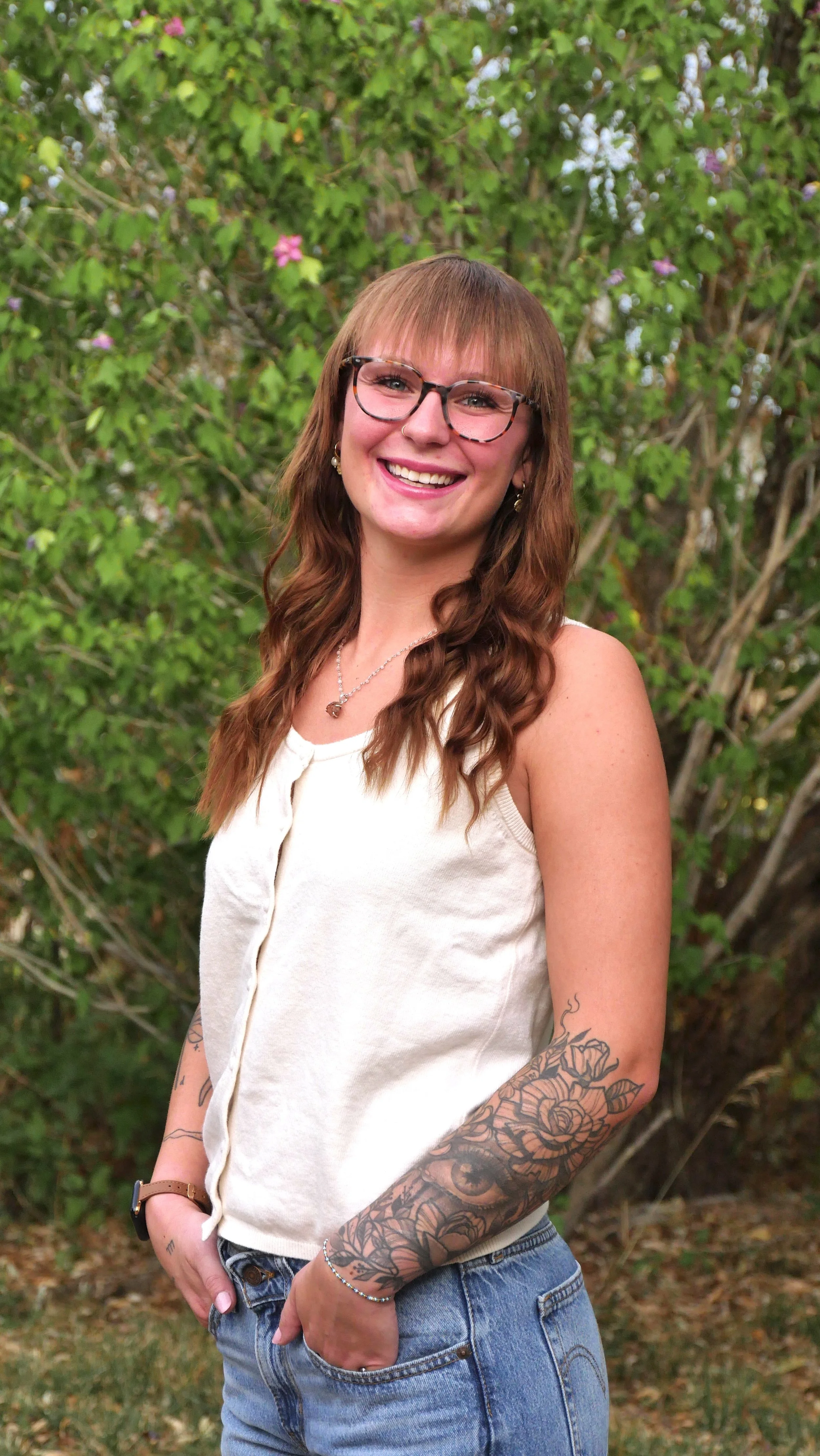 A young woman with long brown hair, glasses, and a nose ring, smiling outdoors in front of green foliage and blooming flowers, wearing a sleeveless beige top and blue jeans, with tattoos on her arms.