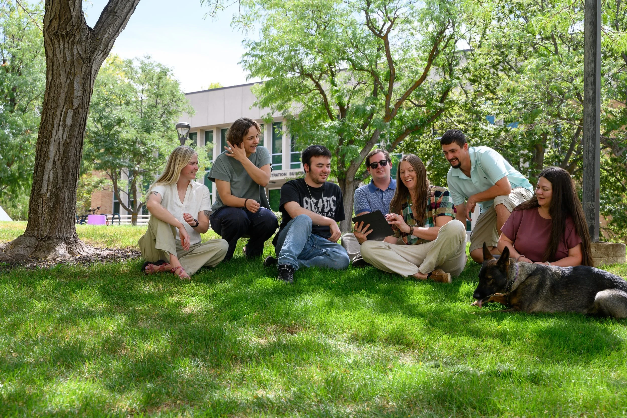 Large group of young adults seated on a lawn under a tree with a dog