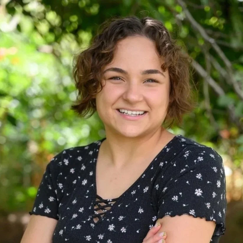 A young woman with short, curly brown hair smiling outdoors with a background of green foliage.