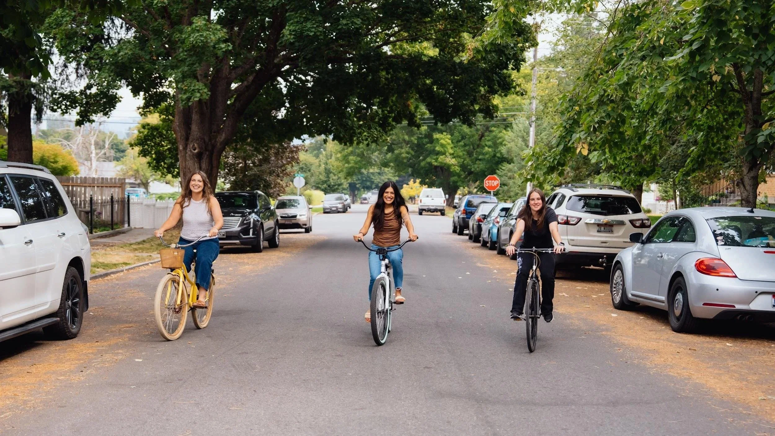 Three women riding bicycles down a residential street lined with parked cars, trees, and a stop sign in the background.