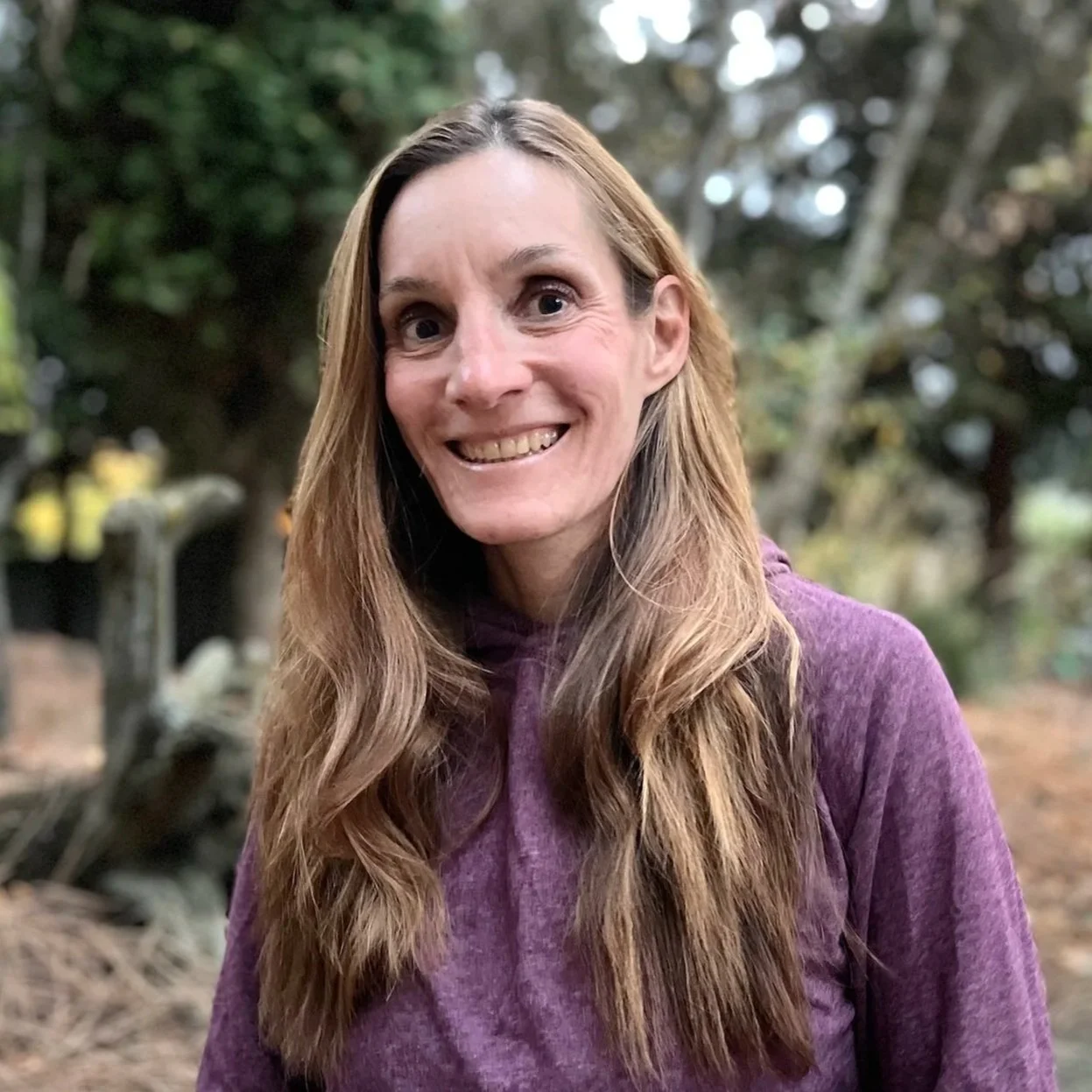 A woman with long, light brown hair, smiling outdoors in a wooded area, wearing a purple hoodie.