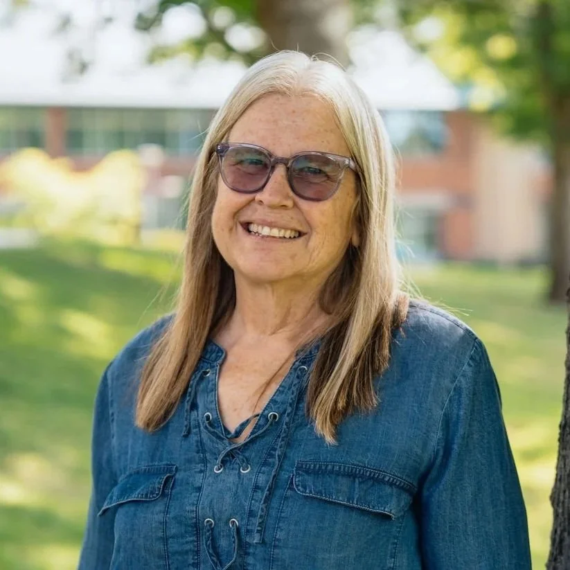 Smiling woman with long blonde hair wearing glasses and a denim shirt outdoors on a sunny day.