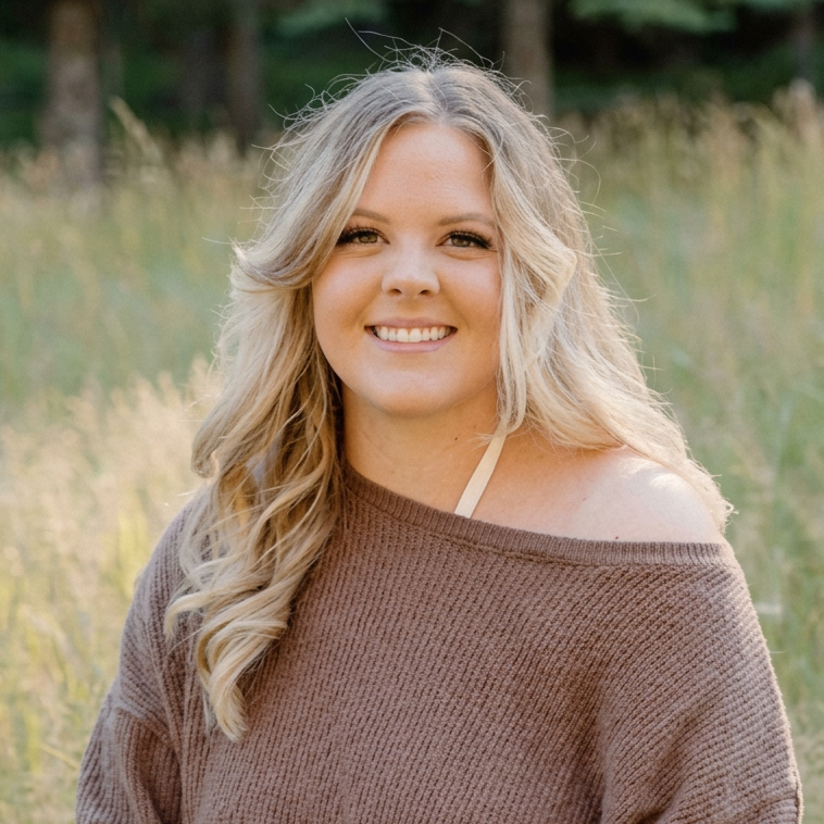 A young woman with long blonde curly hair smiling outdoors in a field, wearing a brown off-shoulder sweater.