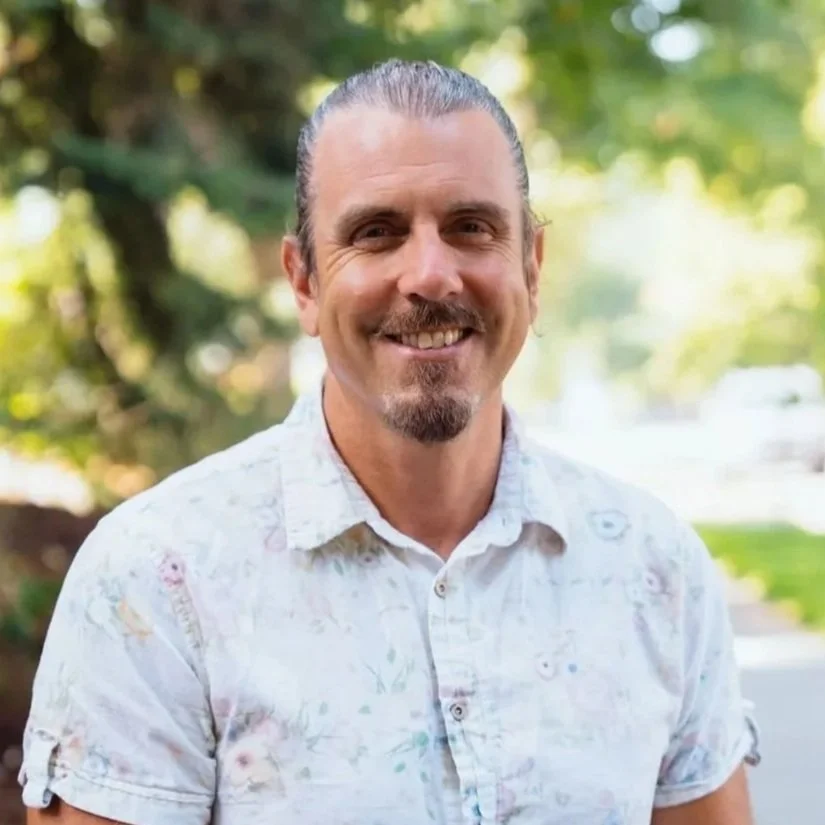 A man with slicked-back hair, a beard and mustache, smiling outdoors with trees and greenery in the background, wearing a light-colored, patterned button-up shirt.