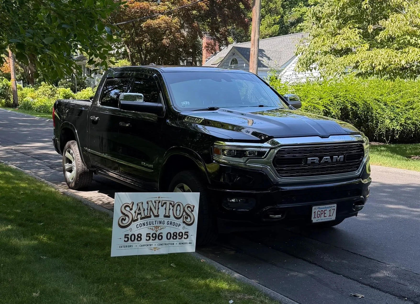 A black RAM pickup truck parked on a suburban street with green trees and houses in the background. A sign in front of the truck displays contact information for Santos Consulting Group.