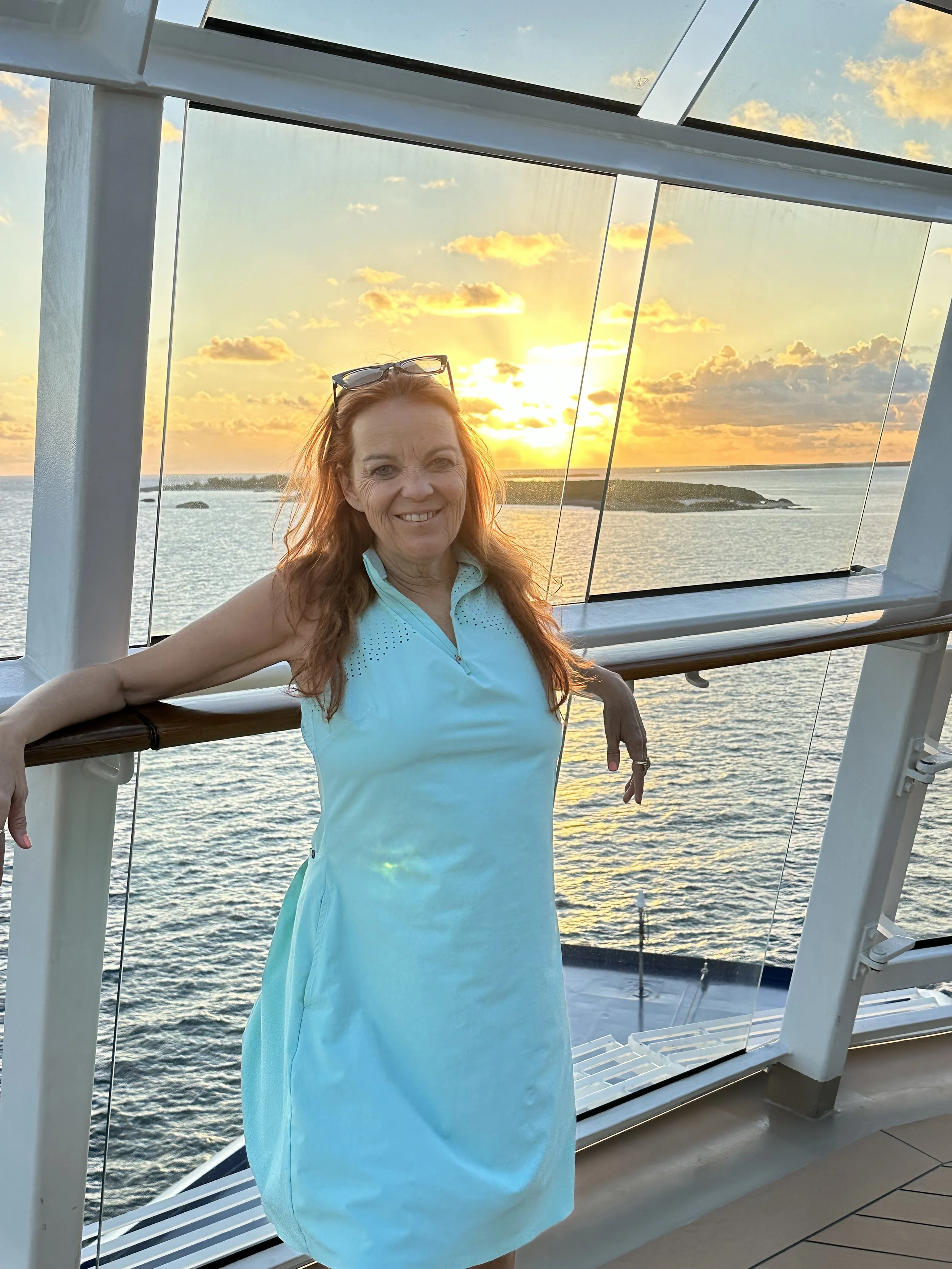 A smiling woman in a light blue dress with sunglasses on her head, standing on a cruise ship deck with a sunset over the ocean in the background.
