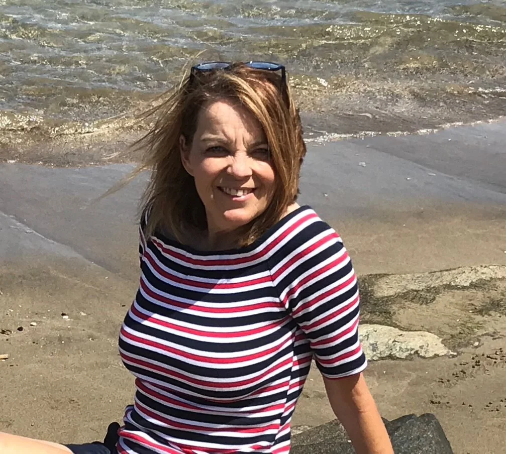 A woman with shoulder-length brown hair, wearing sunglasses on her head and a striped shirt, sitting on a sandy beach near the water and smiling at the camera.
