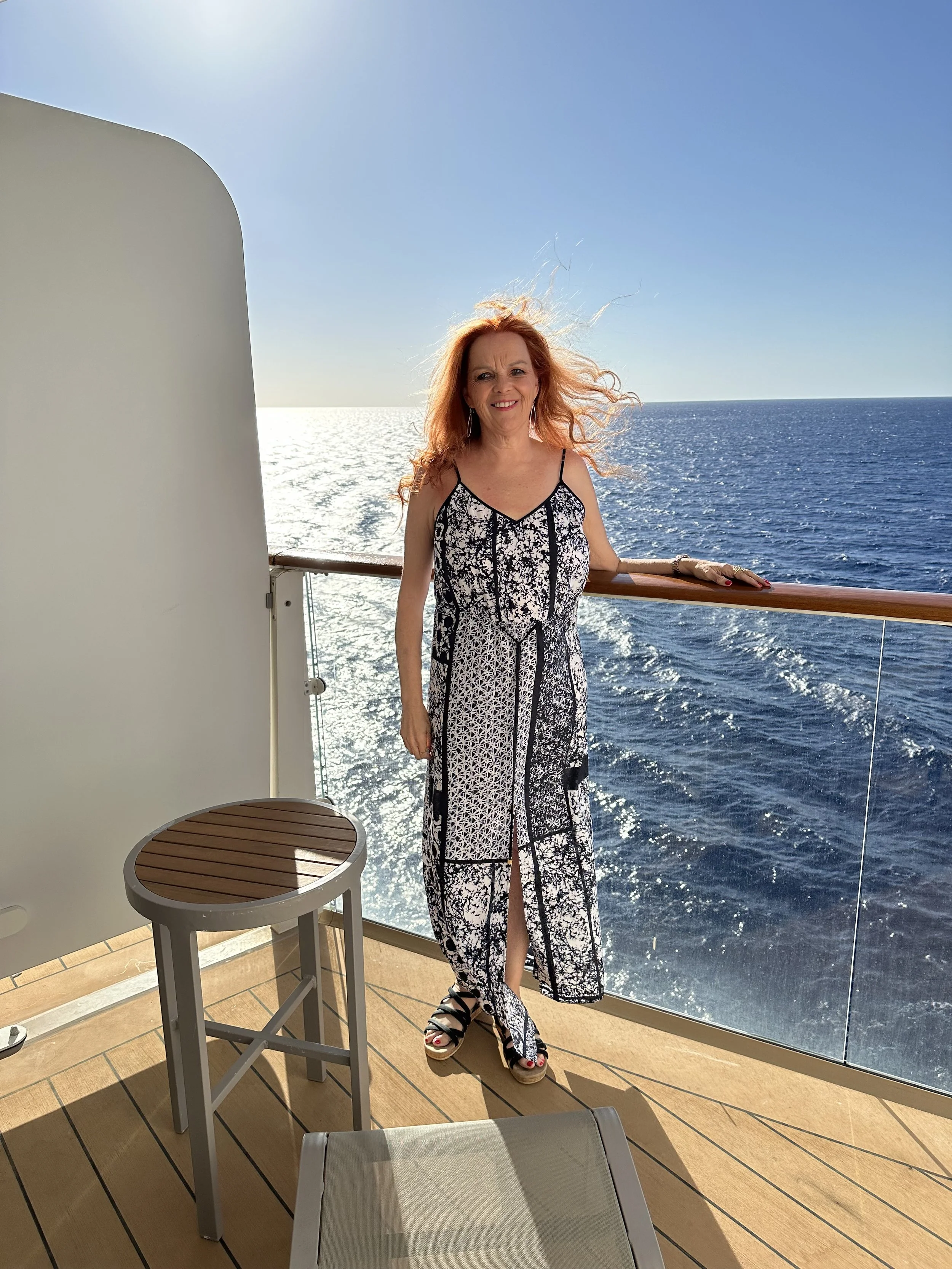 A woman with red hair standing on a ship's deck, smiling at the camera, with ocean and clear blue sky in the background.