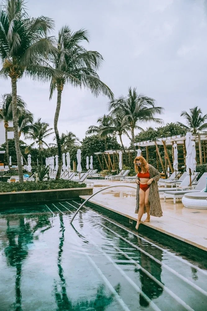 A woman in a red bikini and a long patterned cover-up standing by the edge of a swimming pool with palm trees and lounge chairs in the background.