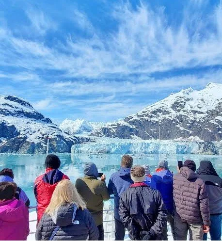 Group of tourists viewing a glacier in a snowy mountain landscape with icy water and partly cloudy sky.