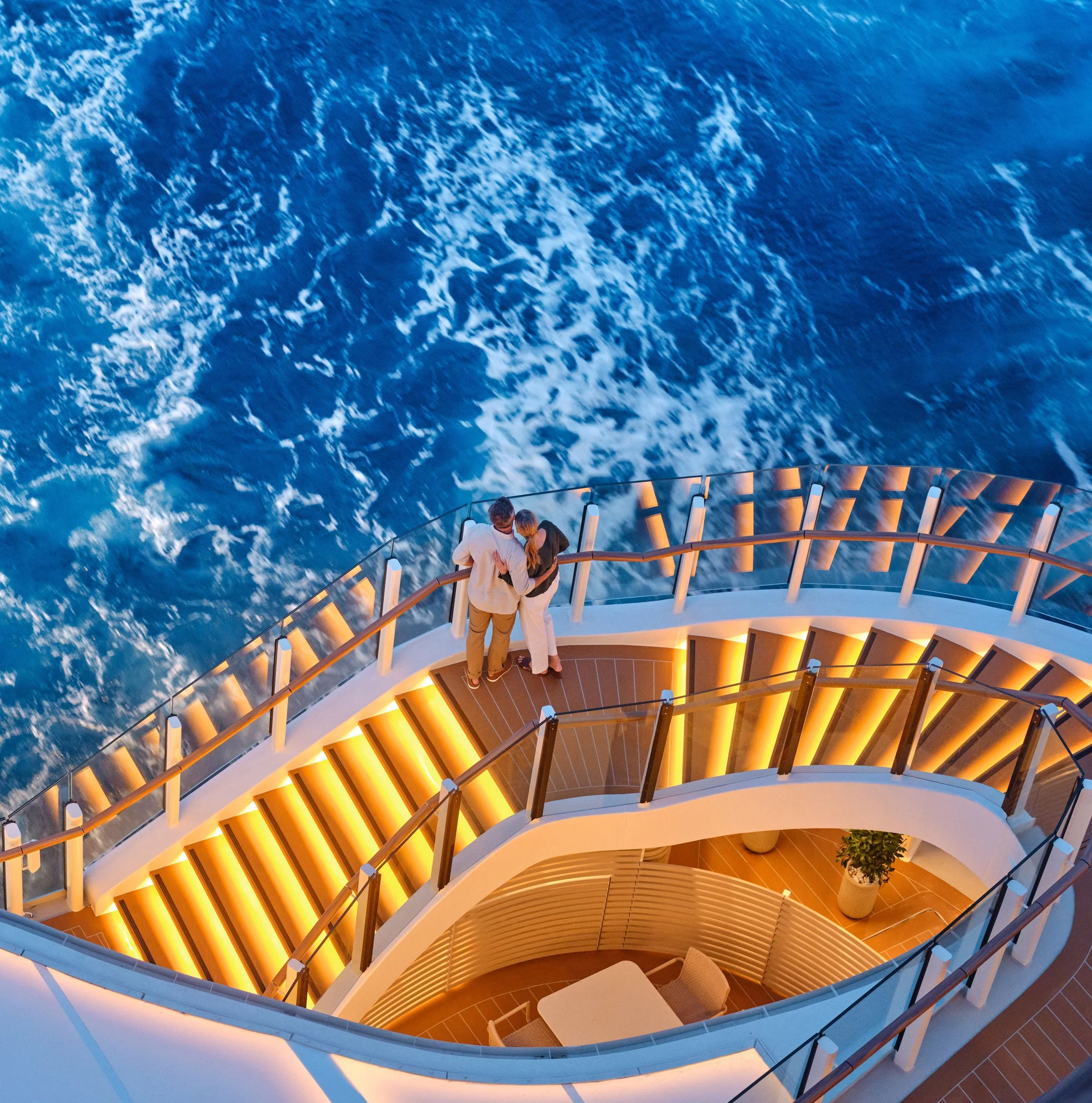 People standing on a curved outdoor stairway overlooking the ocean at sunset, with clouds and the horizon visible in the distance.