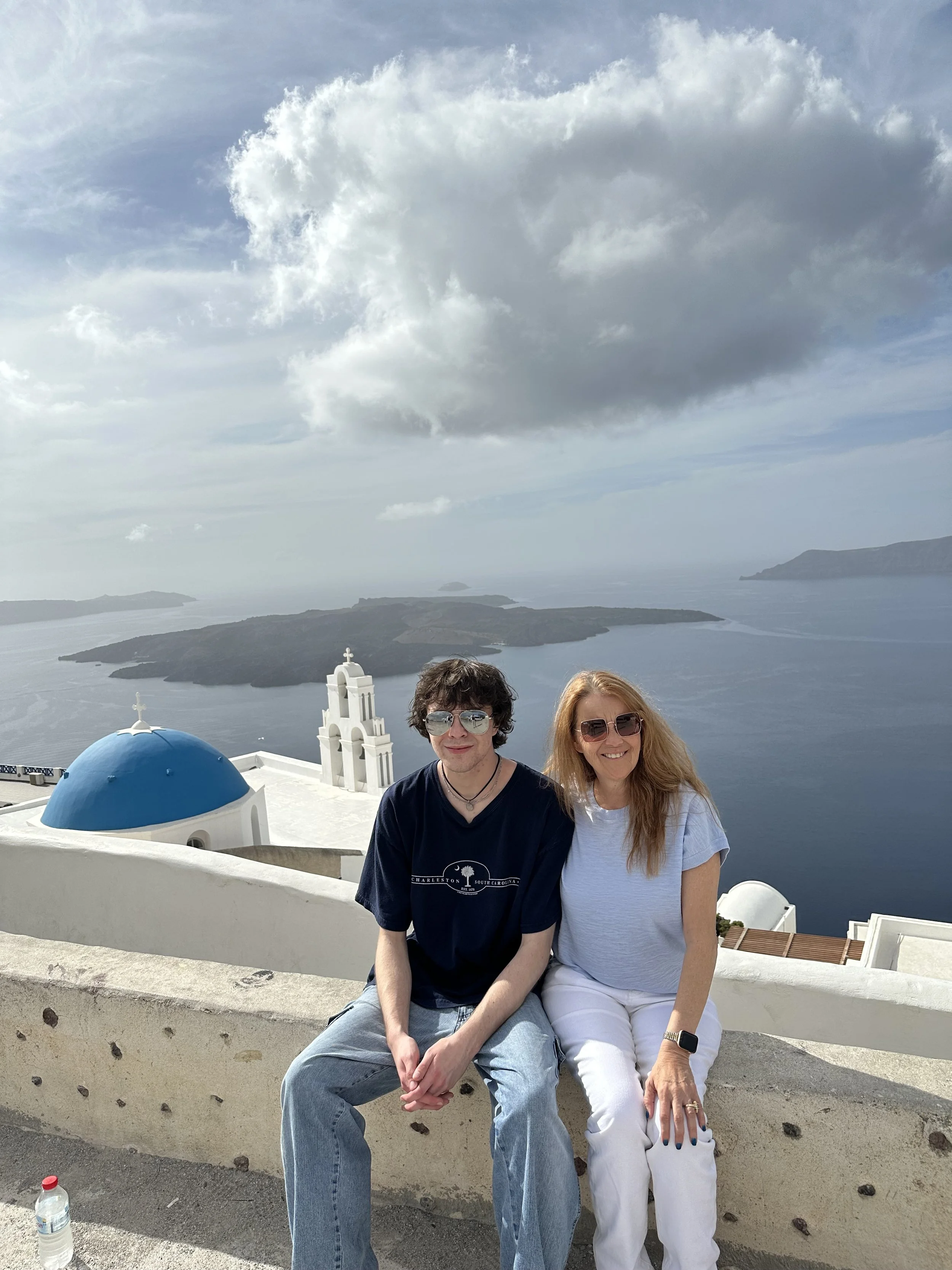 Two people, a man and a woman, sitting on a white wall in front of a scenic view of islands, water, and sky with clouds. The scene suggests a coastal location, possibly Santorini, Greece.