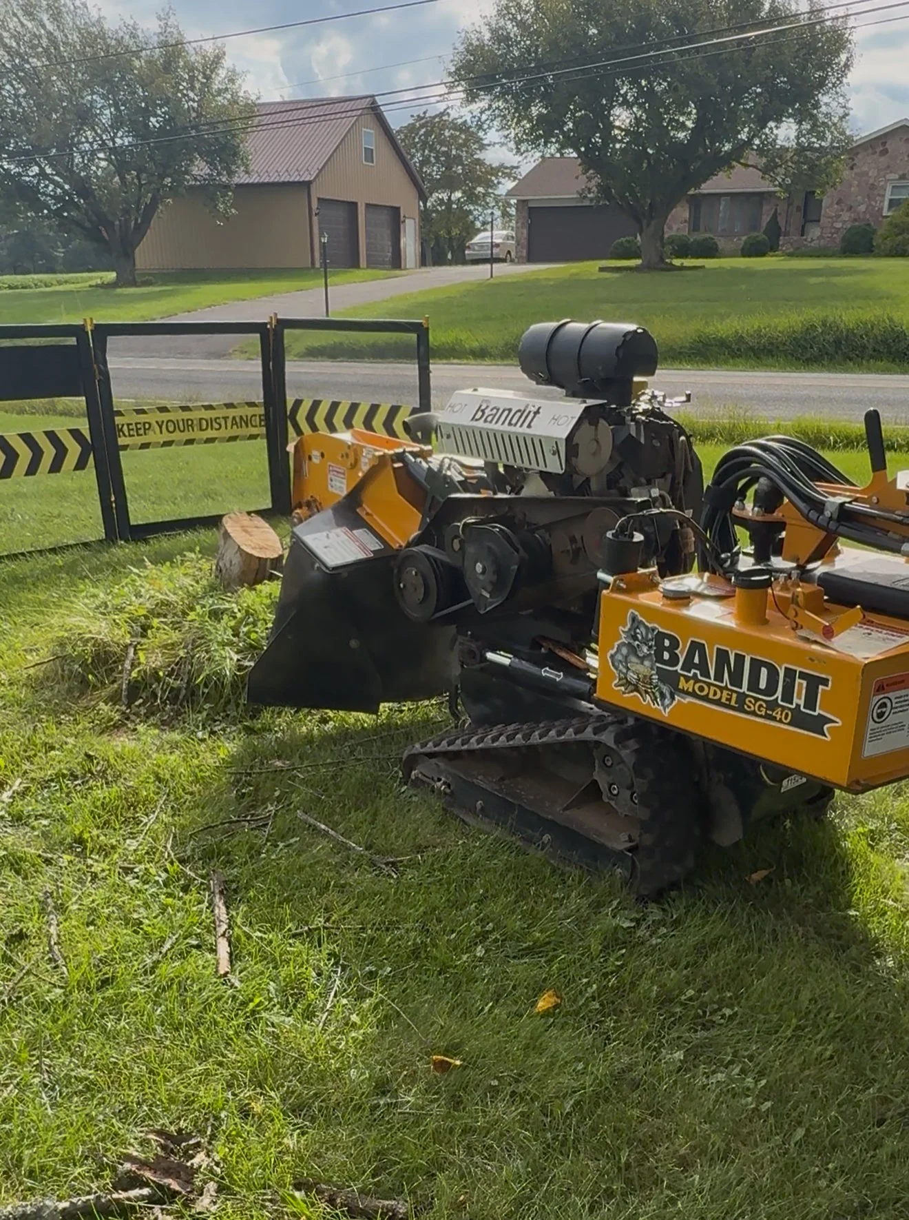 A small, tracked, yellow and black wood cutting machine labeled 'Bandit Model SG-40' on a grassy lawn near a black and yellow caution fence with 'Keep Your Distance' warning, with houses, trees, and a driveway in the background.