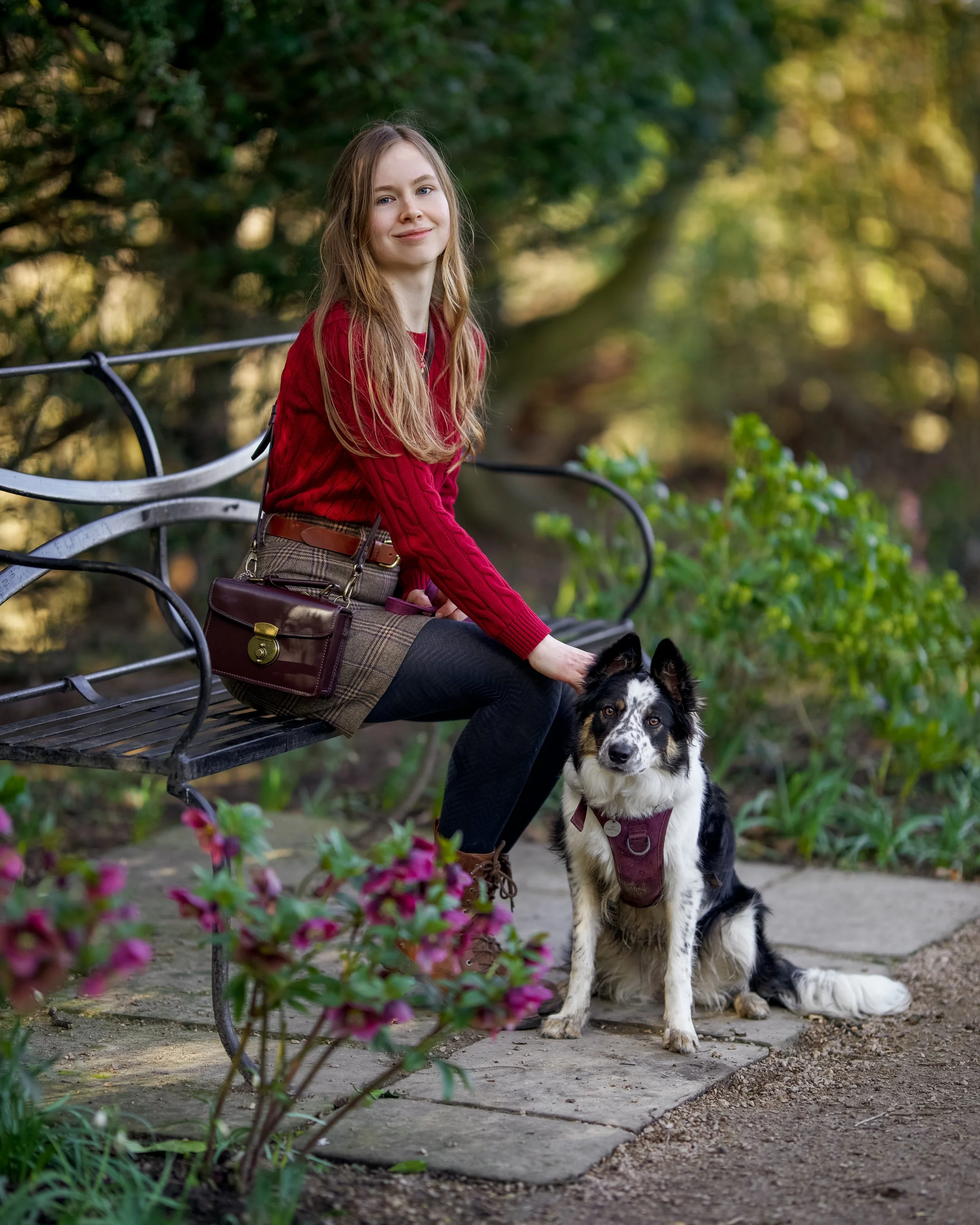 A young woman with long light brown hair sitting on a black park bench in a park, smiling, with a black and white Border Collie dog with a maroon harness sitting next to her. The background features lush greenery and pink flowers in the foreground.