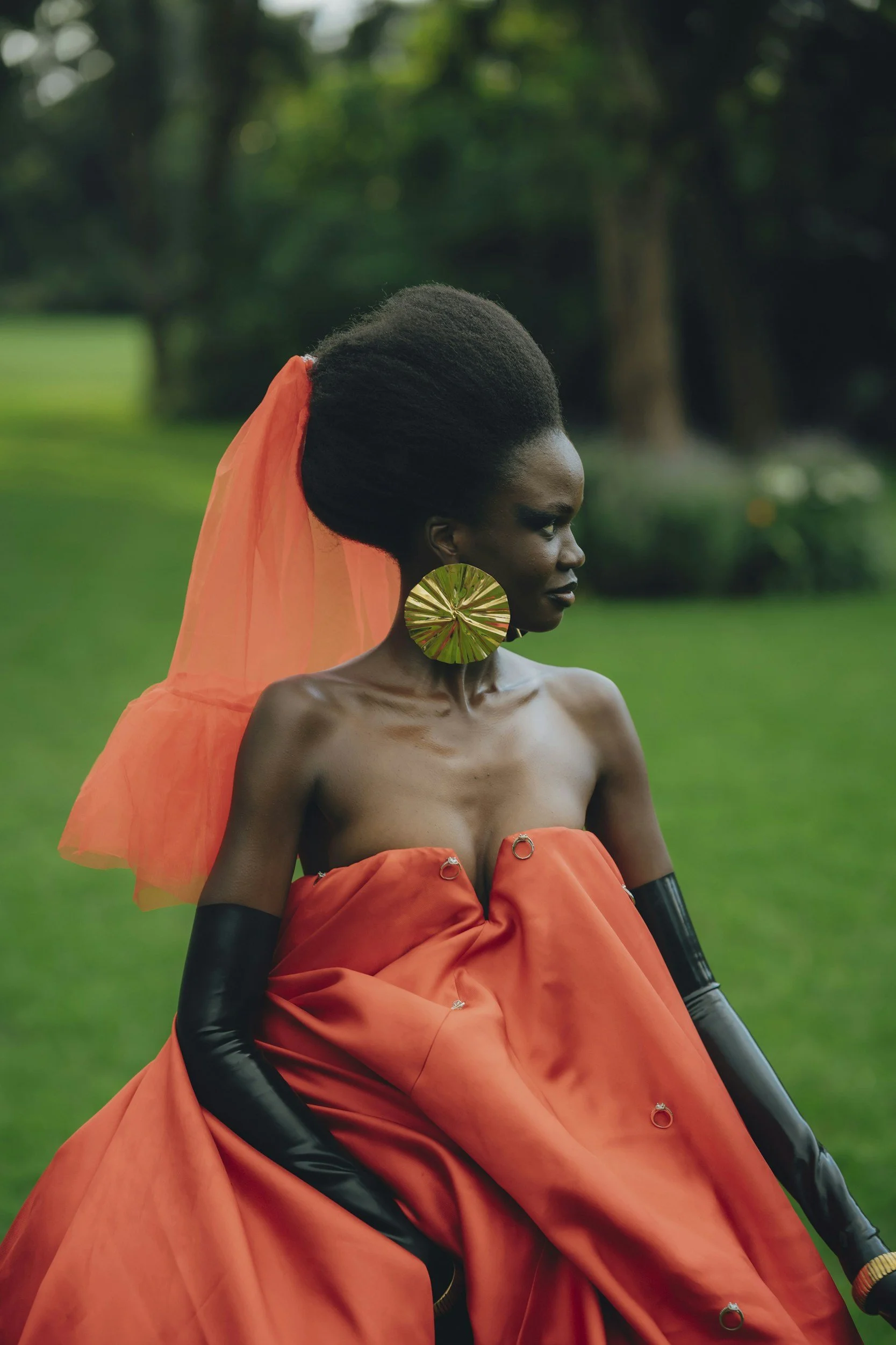 A woman with dark skin and natural hair styled in an afro, adorned with a large gold circular earring, wears an off-shoulder orange dress with a matching orange veil or fabric. She stands outdoors against a blurred green natural background, looking to her right.