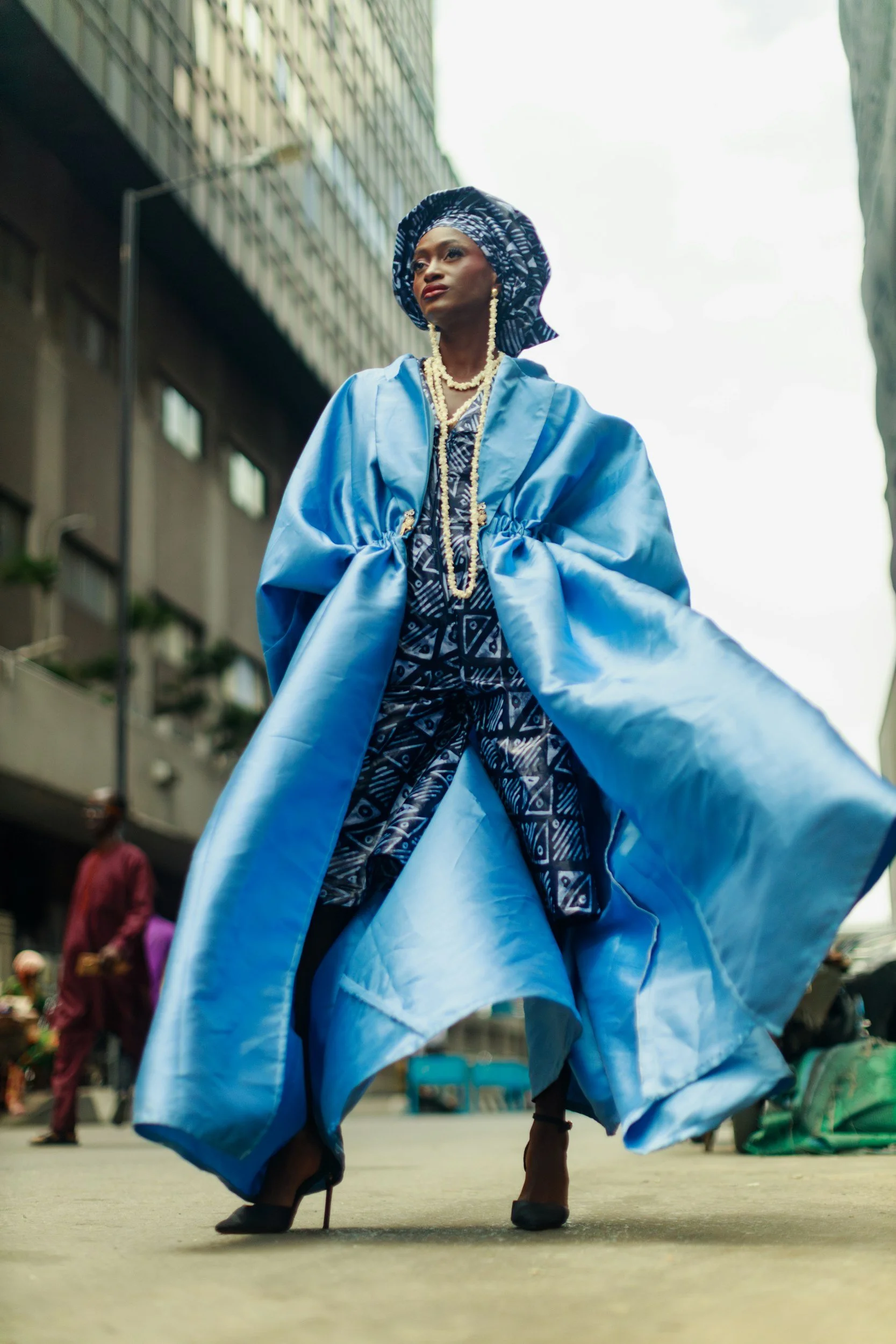 A woman dressed in traditional African attire, featuring a headwrap and a flowing blue garment, is walking confidently on a city street, with buildings and pedestrians in the background.