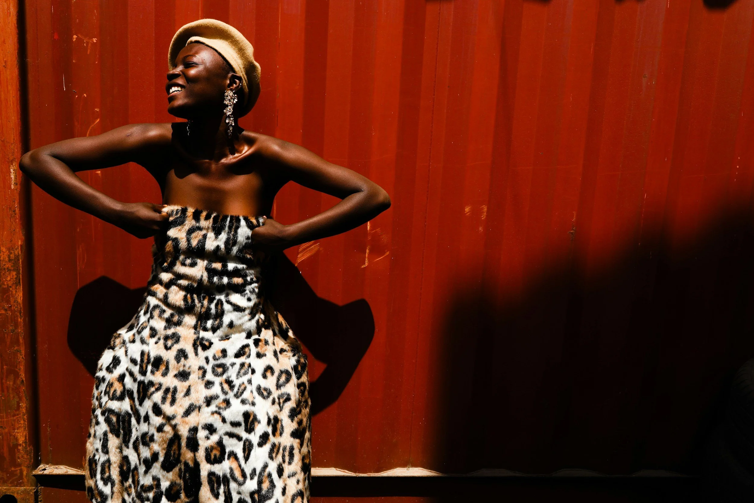 A woman dressed in leopard print fabric poses against a rusty red metal wall, smiling with her eyes closed, wearing large sparkling earrings, a beige hat, and her arms are crossed on her waist.