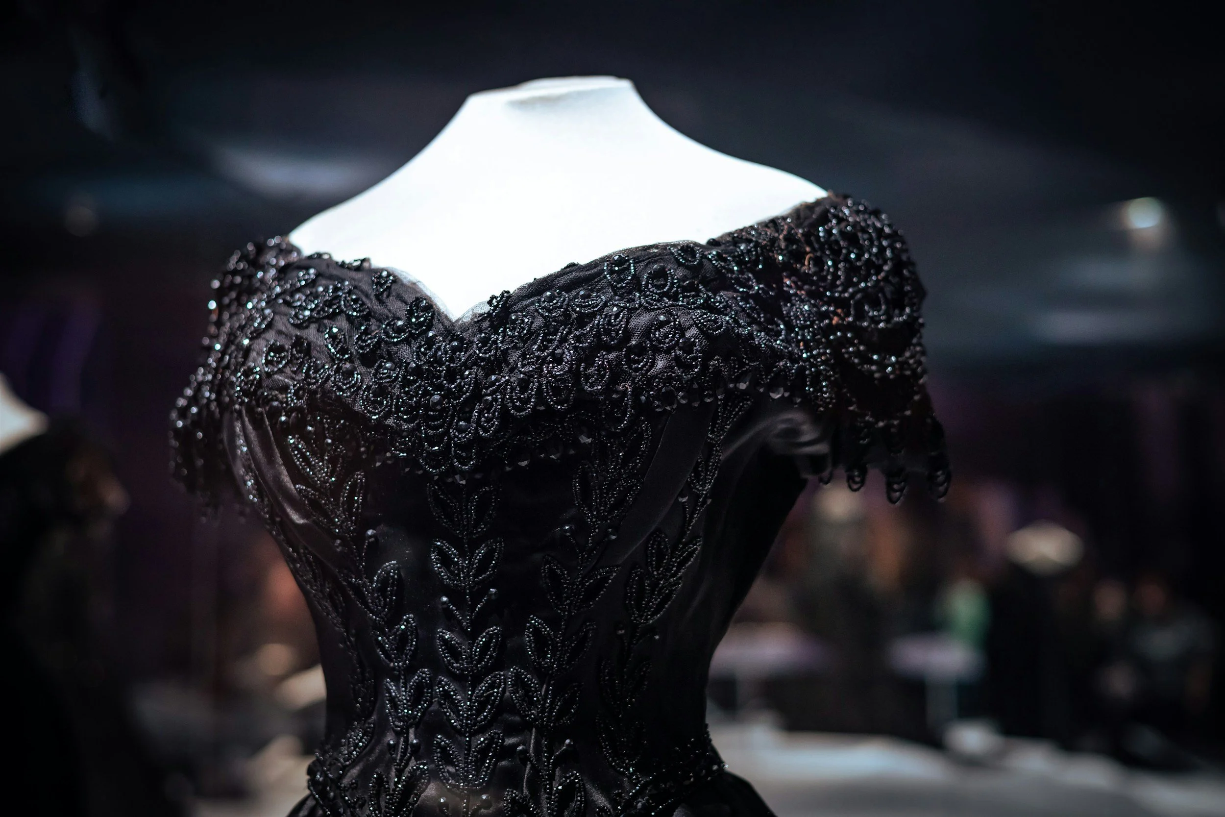 Close-up of a black dress with intricate embroidery and beadwork, displayed on a mannequin in a dark setting.