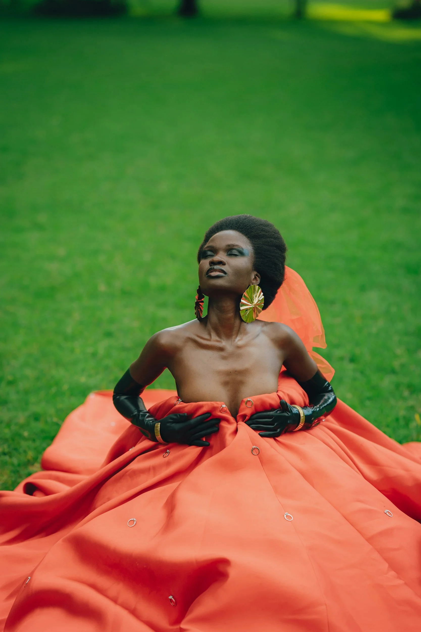 A woman with dark skin and an elaborate afro hairstyle sits on a lush green lawn. She wears large, colorful earrings and a vivid orange, voluminous skirt with metal rings. She poses with her eyes closed and hands on her abdomen, exuding confidence and grace.