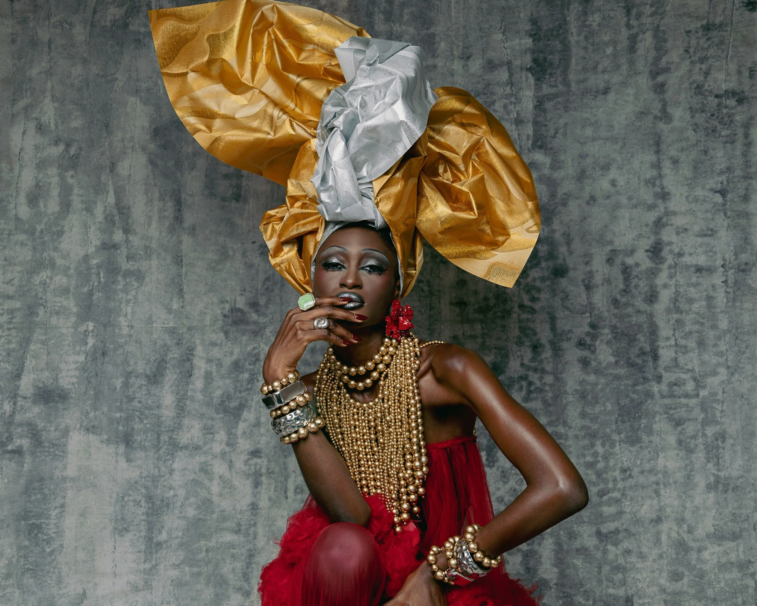 A woman with dark skin wearing an elaborate gold and silver headwrap, red dress, and multiple pearl and silver jewelry posed against a gray textured background.