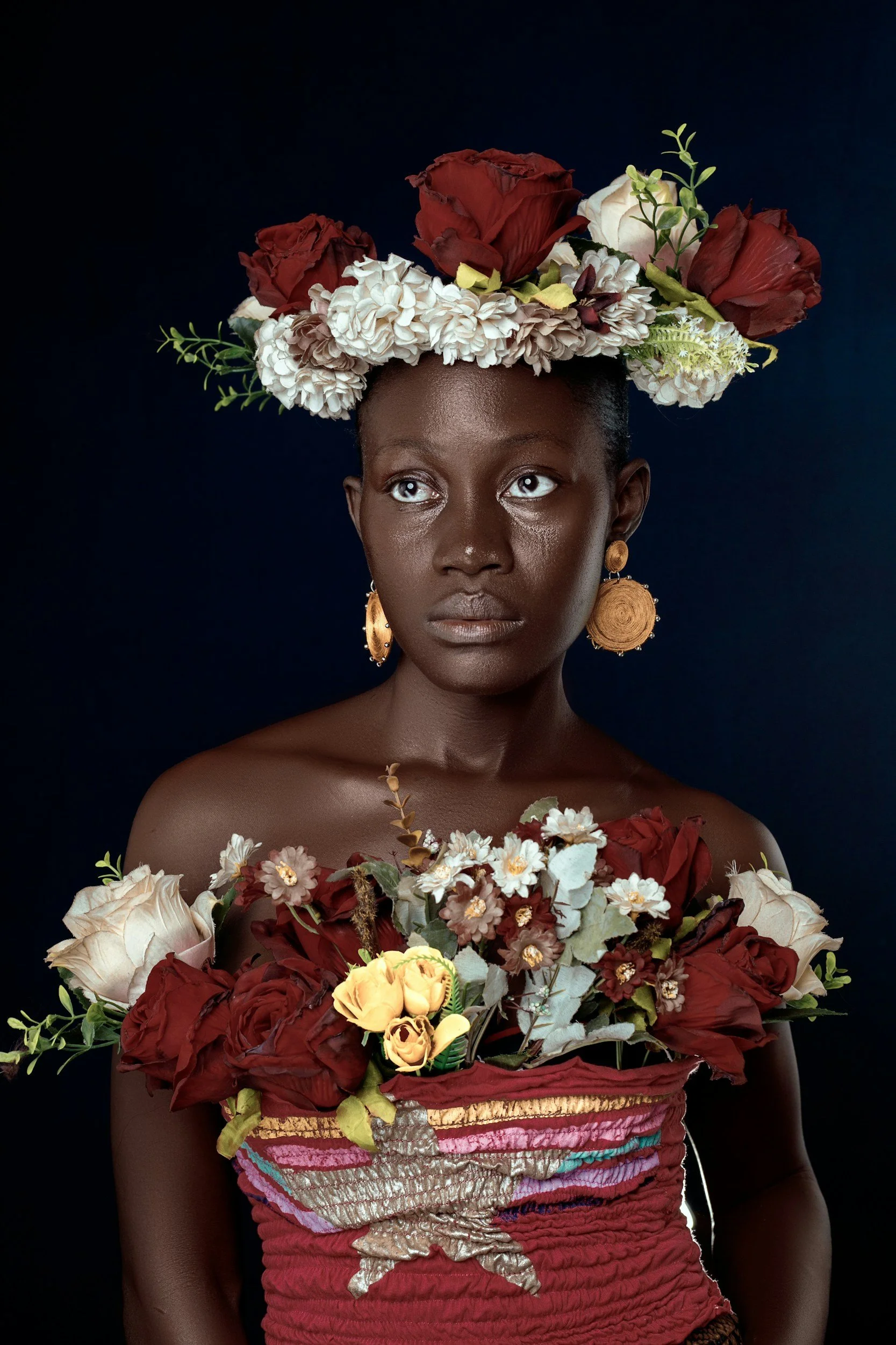 A woman with dark skin, short dark hair, and large gold earrings, wearing a floral crown made of red and white flowers. She holds a large bouquet of flowers in front of her chest. The background is dark blue.