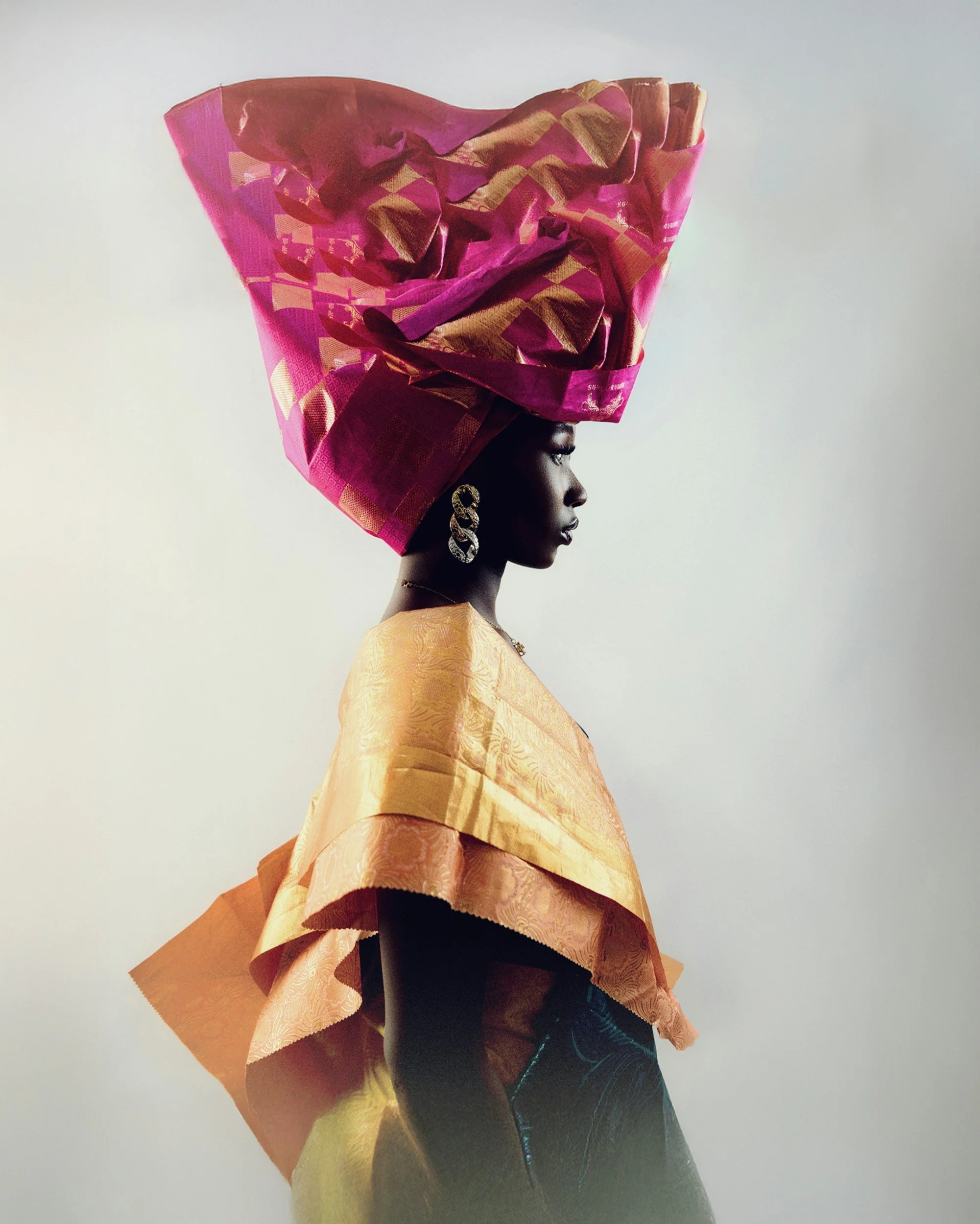 Profile of an African woman wearing a large pink and gold headwrap, elegant earrings, and a patterned yellow and black garment.