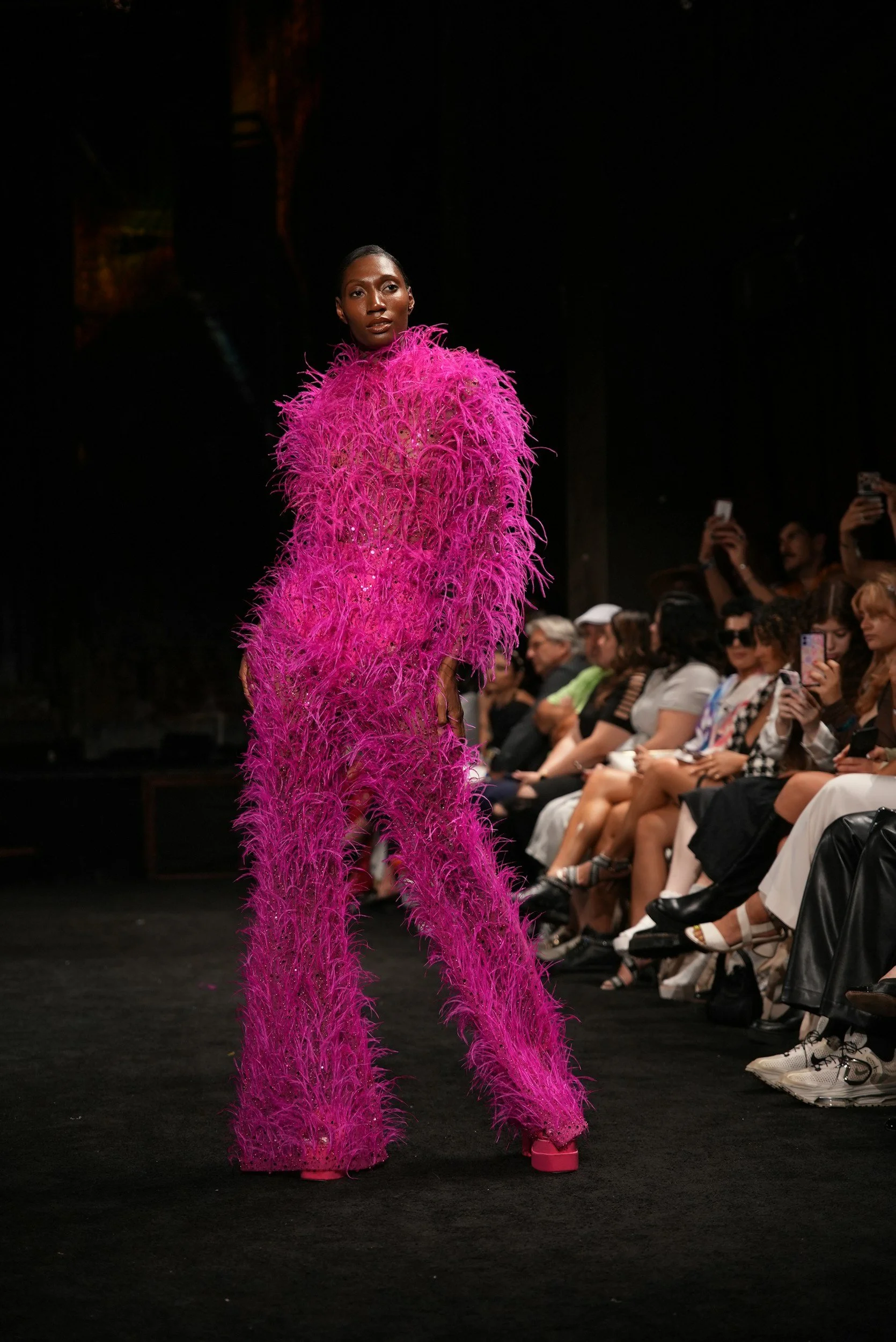 A model walking down the runway wearing a vibrant, pink, feathered outfit during a fashion show, with an audience taking photos in the background.