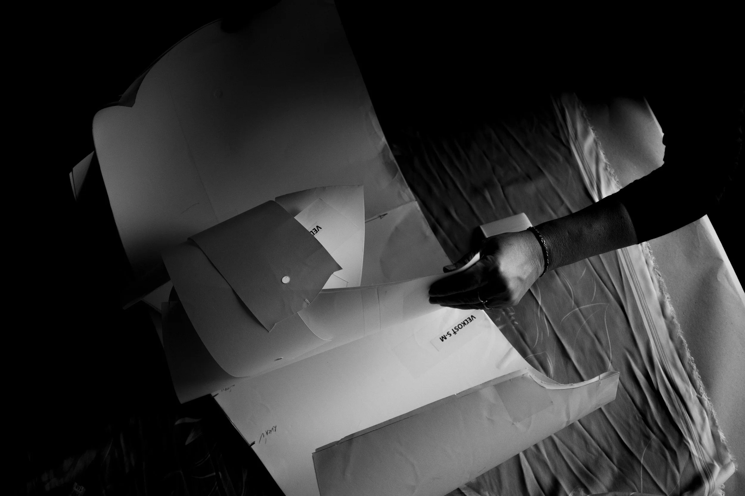 A person wearing a bracelet stacking papers, with one hand holding and another writing or pointing, on a wooden floor. There are boxes or packages nearby.