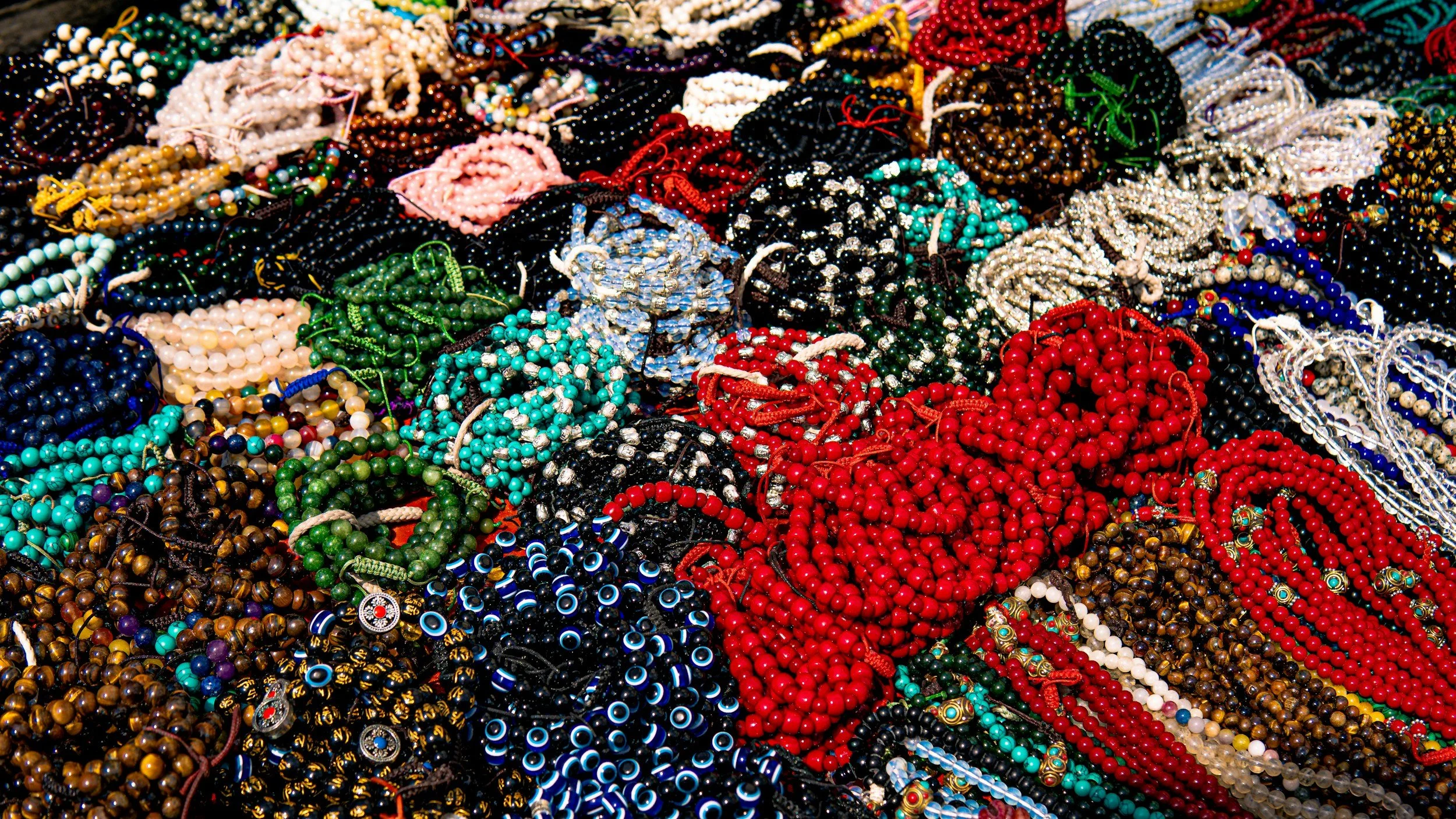 A large collection of colorful beaded necklaces and bracelets piled together. Various colors including red, blue, green, black, white, pink, and brown, with different bead sizes and patterns.
