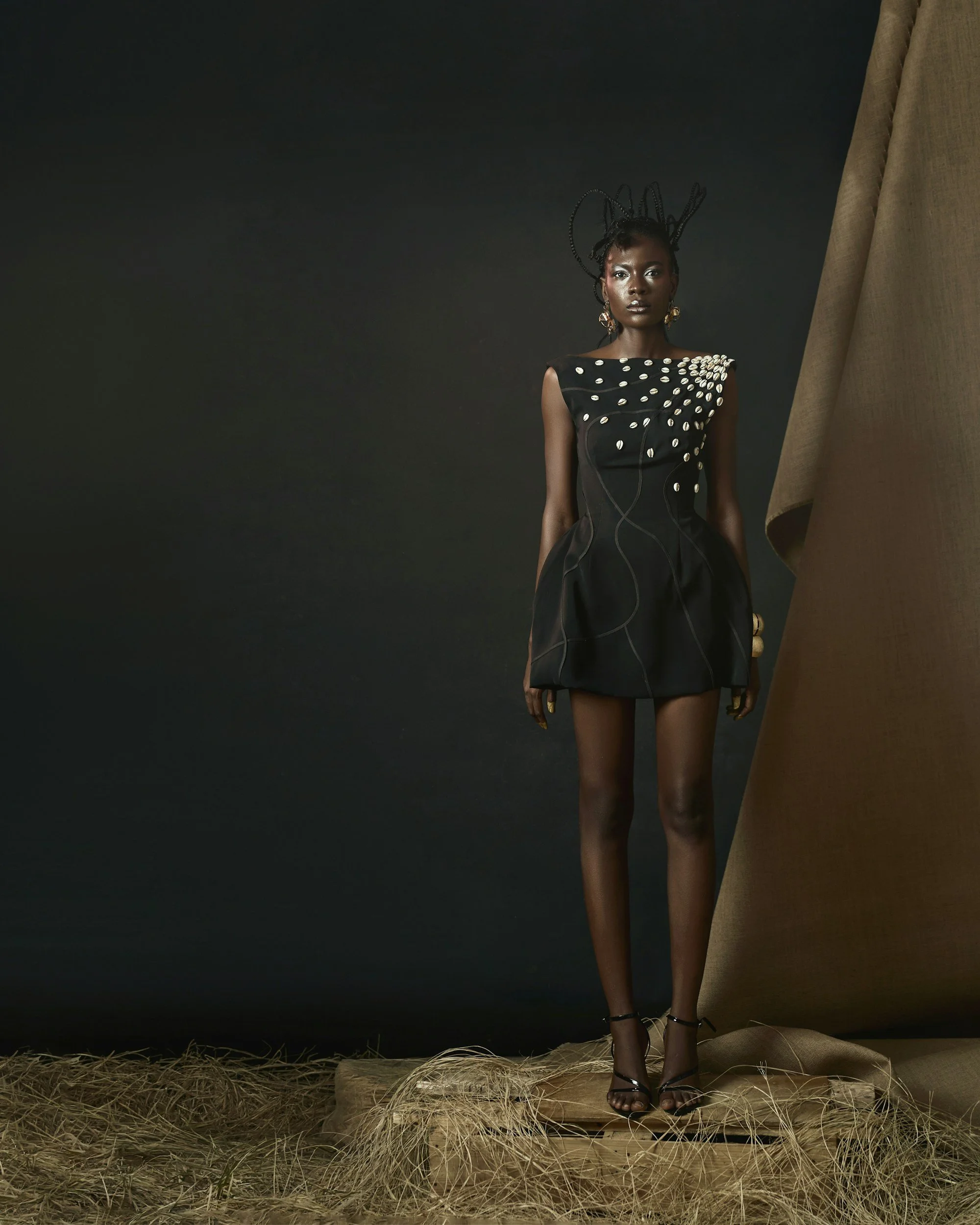 A woman with dark skin and braided hair wearing a black dress decorated with white shell-like accessories, standing on a wooden platform with straw underneath, against a dark background.