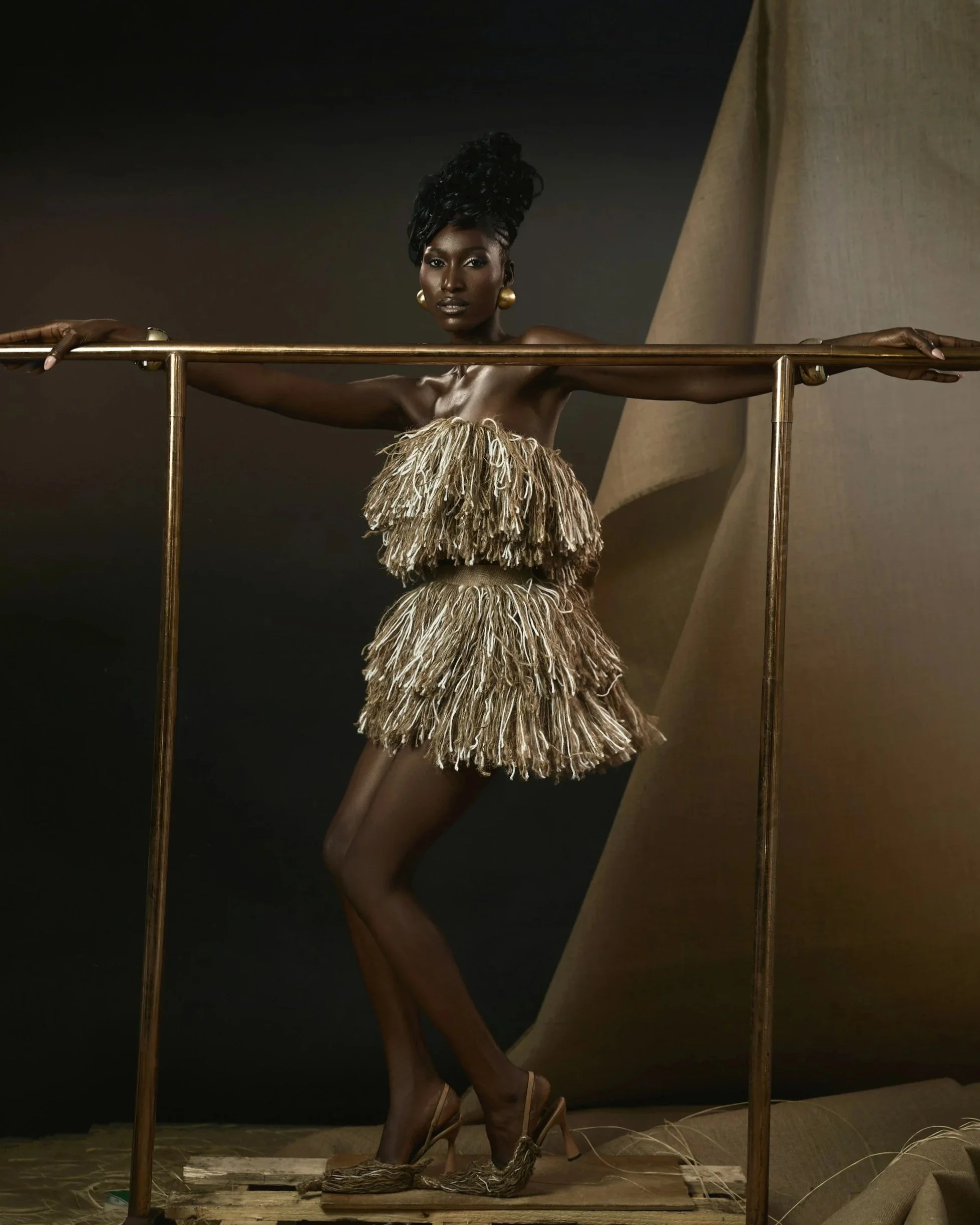 A woman with dark skin and an elaborate braided hairstyle poses behind a brass railing, wearing a strapless dress made of textured, fringed fabric, with high-heeled sandals, set against a dark background with a beige drape.