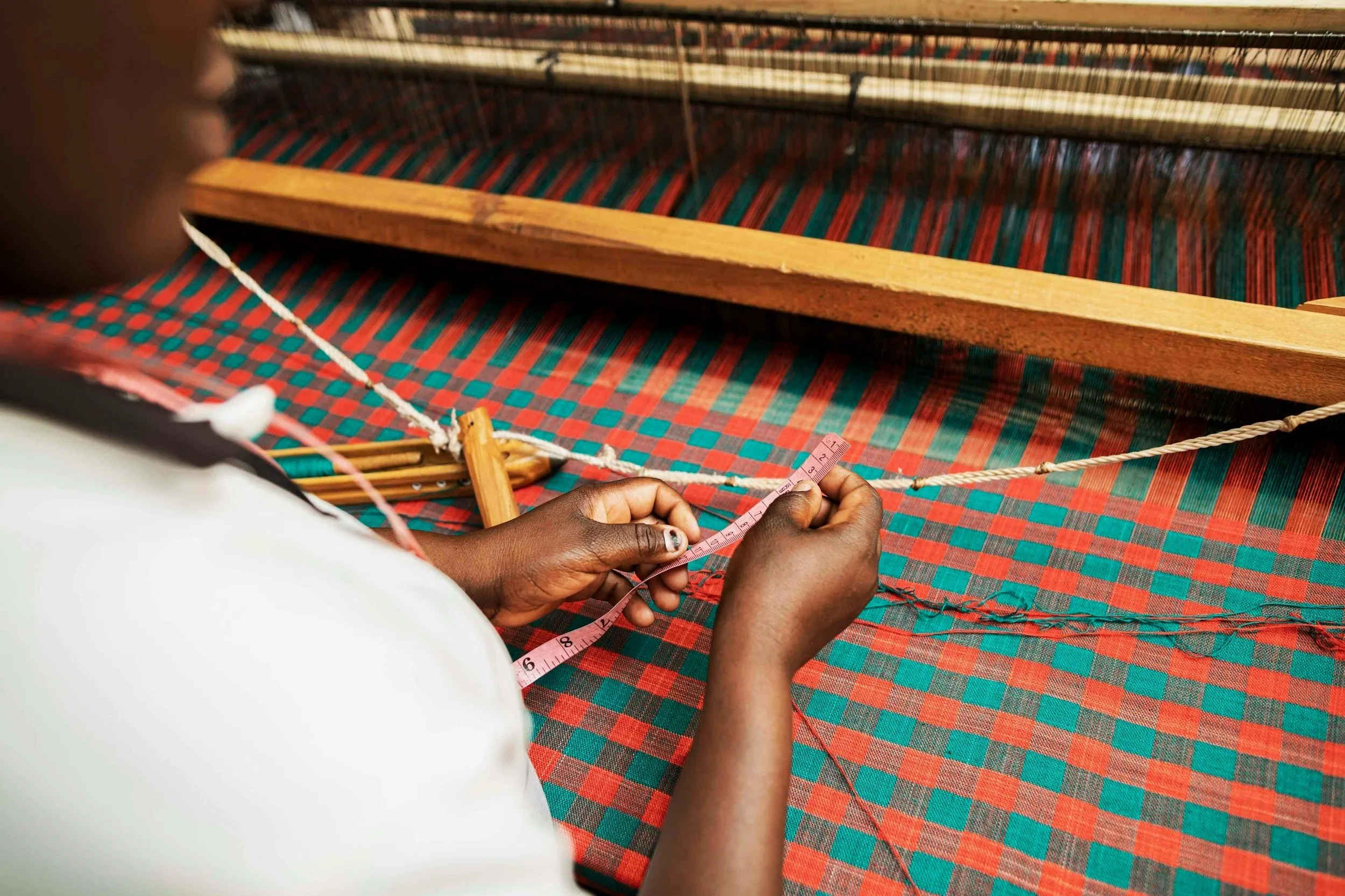 A person measuring a woven fabric on a loom with a pink measuring tape. The fabric has a red and green checkered pattern.