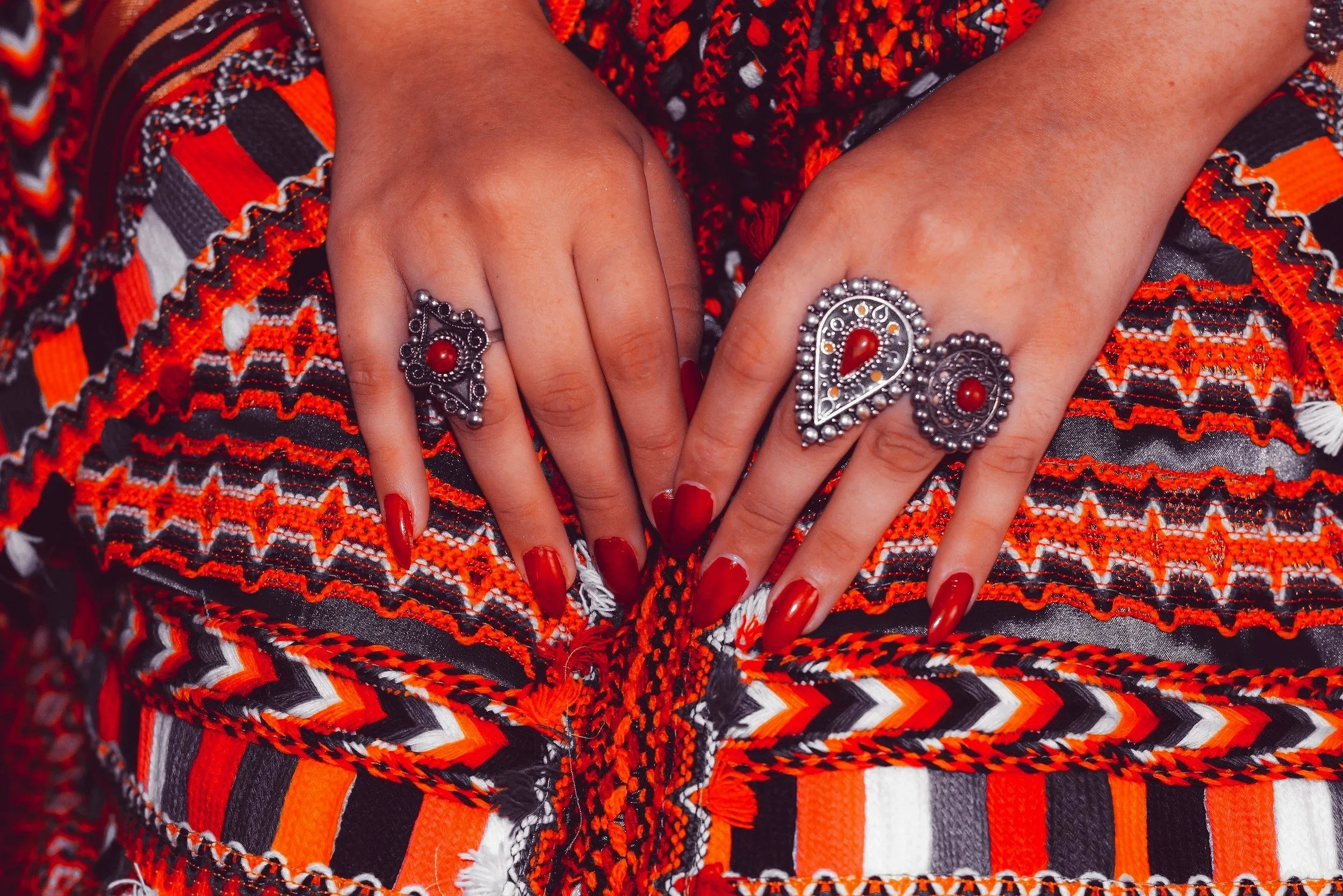 Close-up of a woman's hands resting on colorful, patterned fabric. She is wearing multiple ornate rings with red stones and red nail polish.