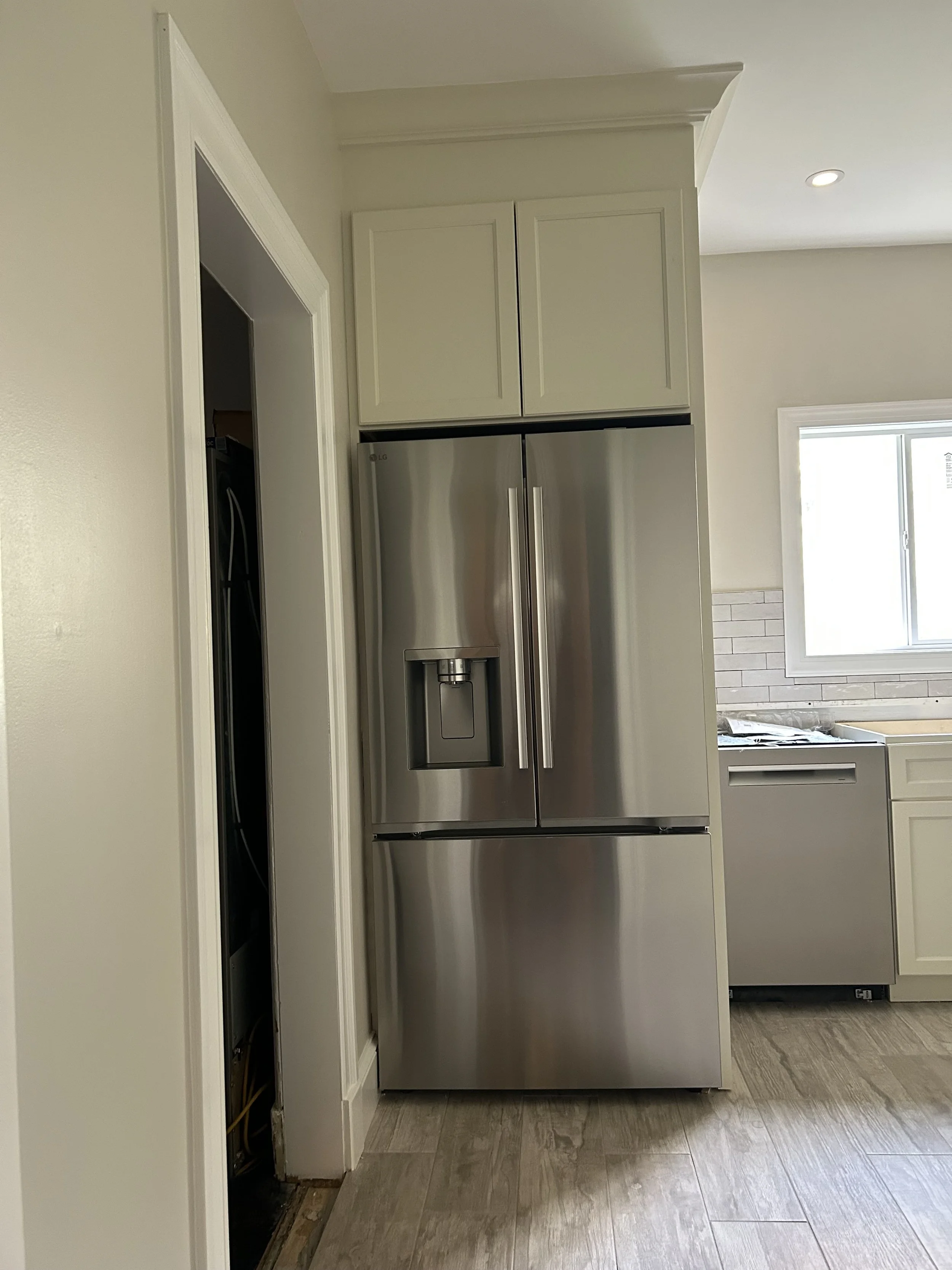 A stainless steel refrigerator in a kitchen with white cabinets, a window, and a wooden floor.