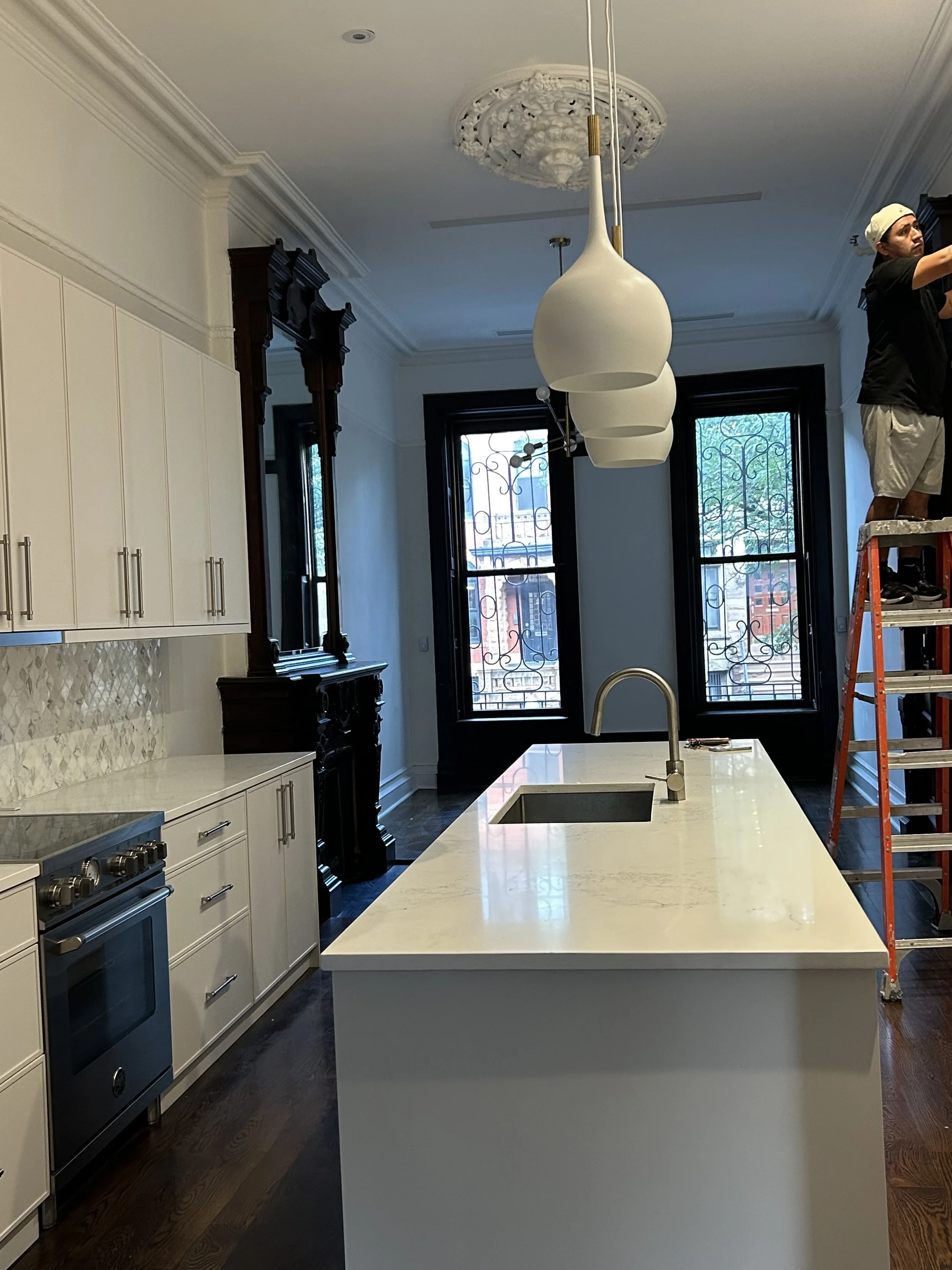 Interior view of a modern kitchen with white cabinets, black trim, a large white kitchen island, large windows with black frames, and a worker on a ladder installing a ceiling light fixture.