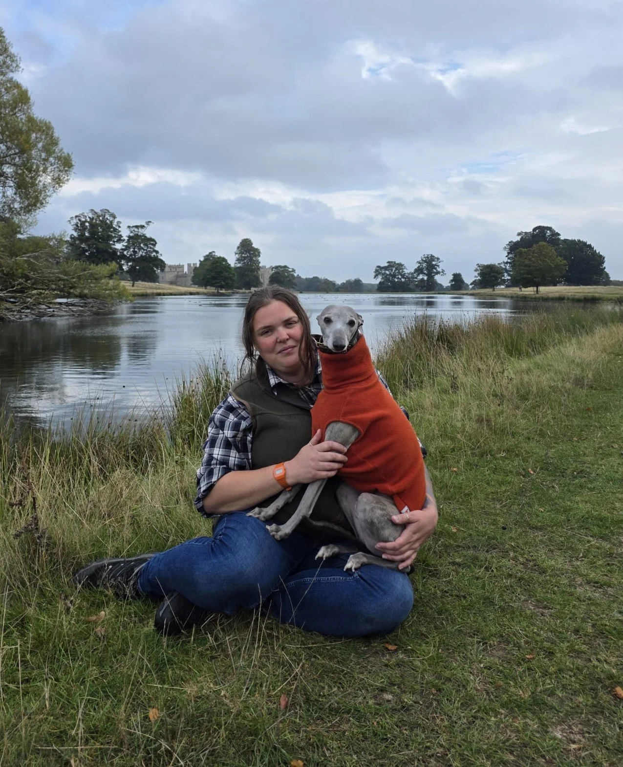 A woman sitting on the grass near a river, holding a greyhound dog in an orange hoodie. The background features a river, trees, and cloudy sky.
