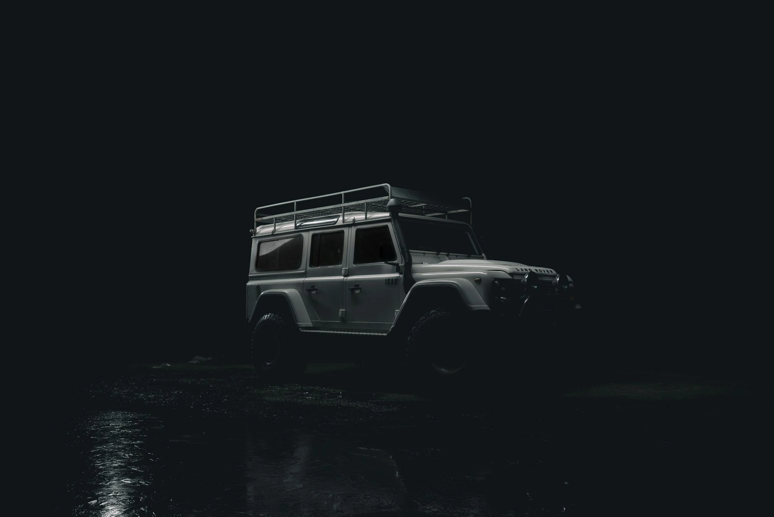 A silver off-road Land Rover vehicle with a roof rack, parked on a wet surface at night, with a dark background.