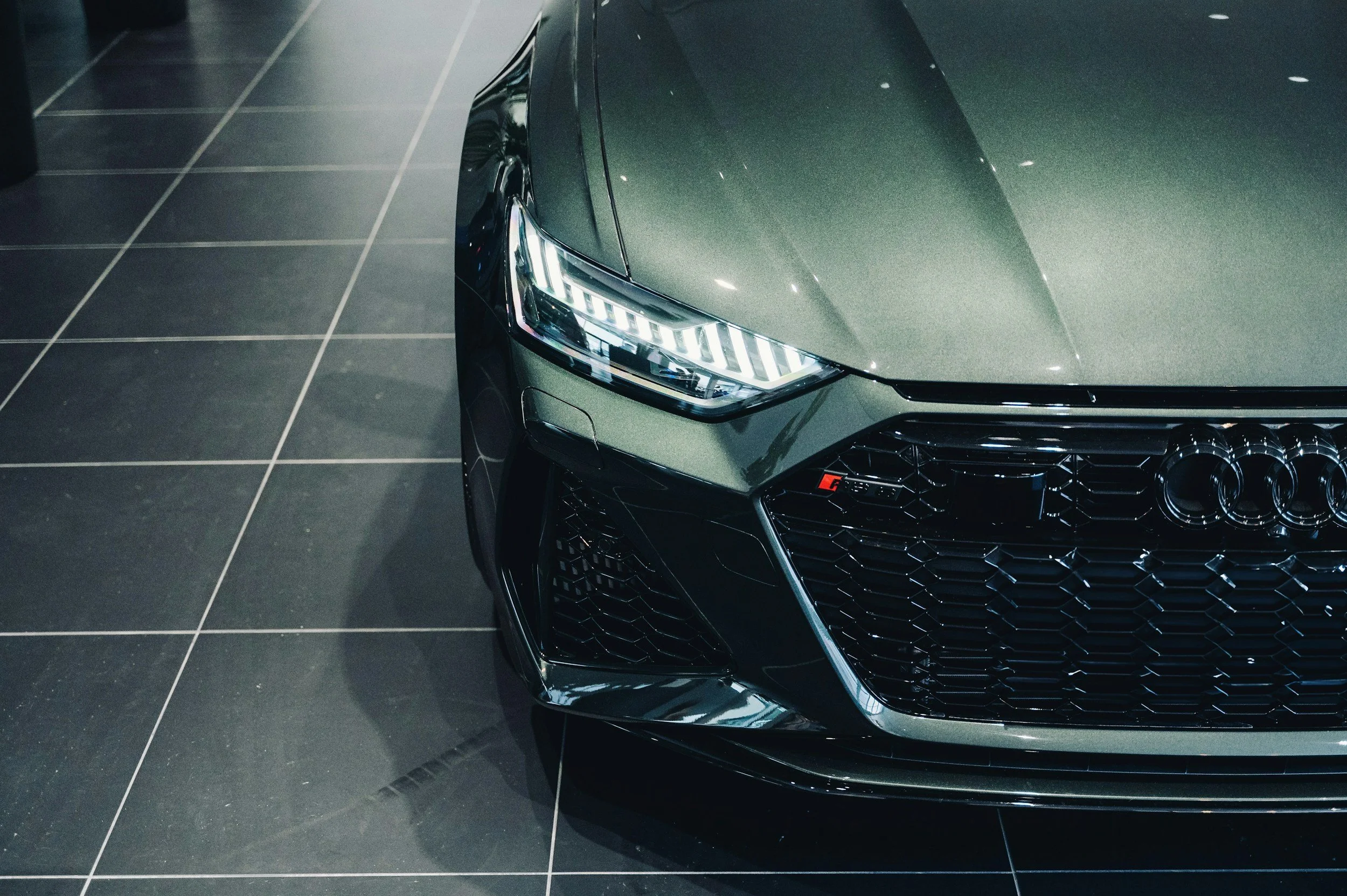 Close-up of the front right side of a dark green Audi car in a showroom, showing the LED headlight, grille, and part of the hood on a tiled floor.