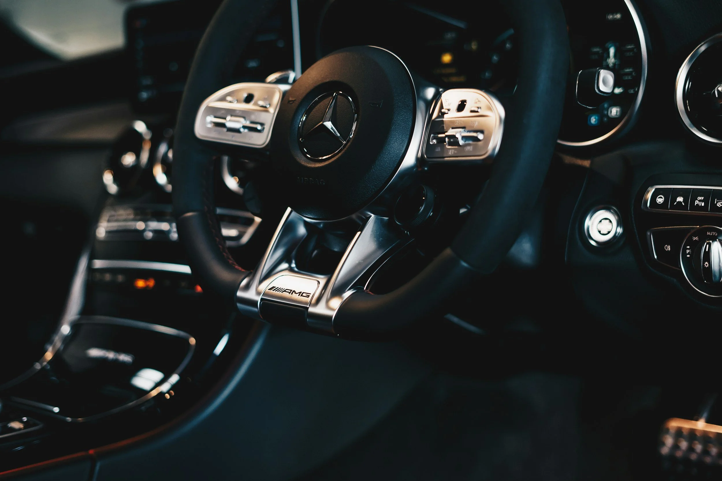 Close-up view of a Mercedes-Benz AMG steering wheel inside a vehicle, showing the Mercedes logo at the center and various control buttons on the sides.