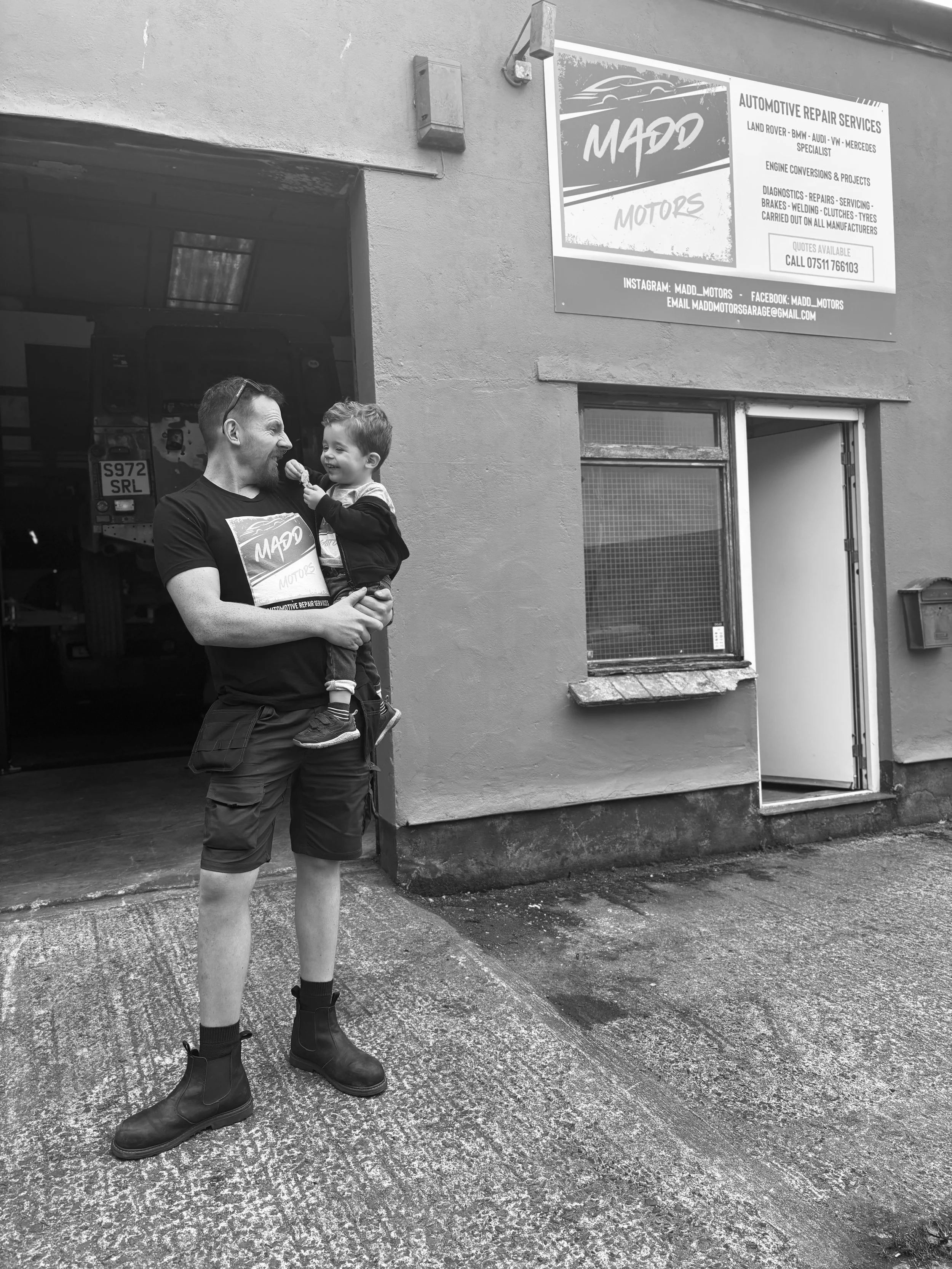 Man holding a smiling child outside a garage, with a sign for an automotive repair shop on the wall.