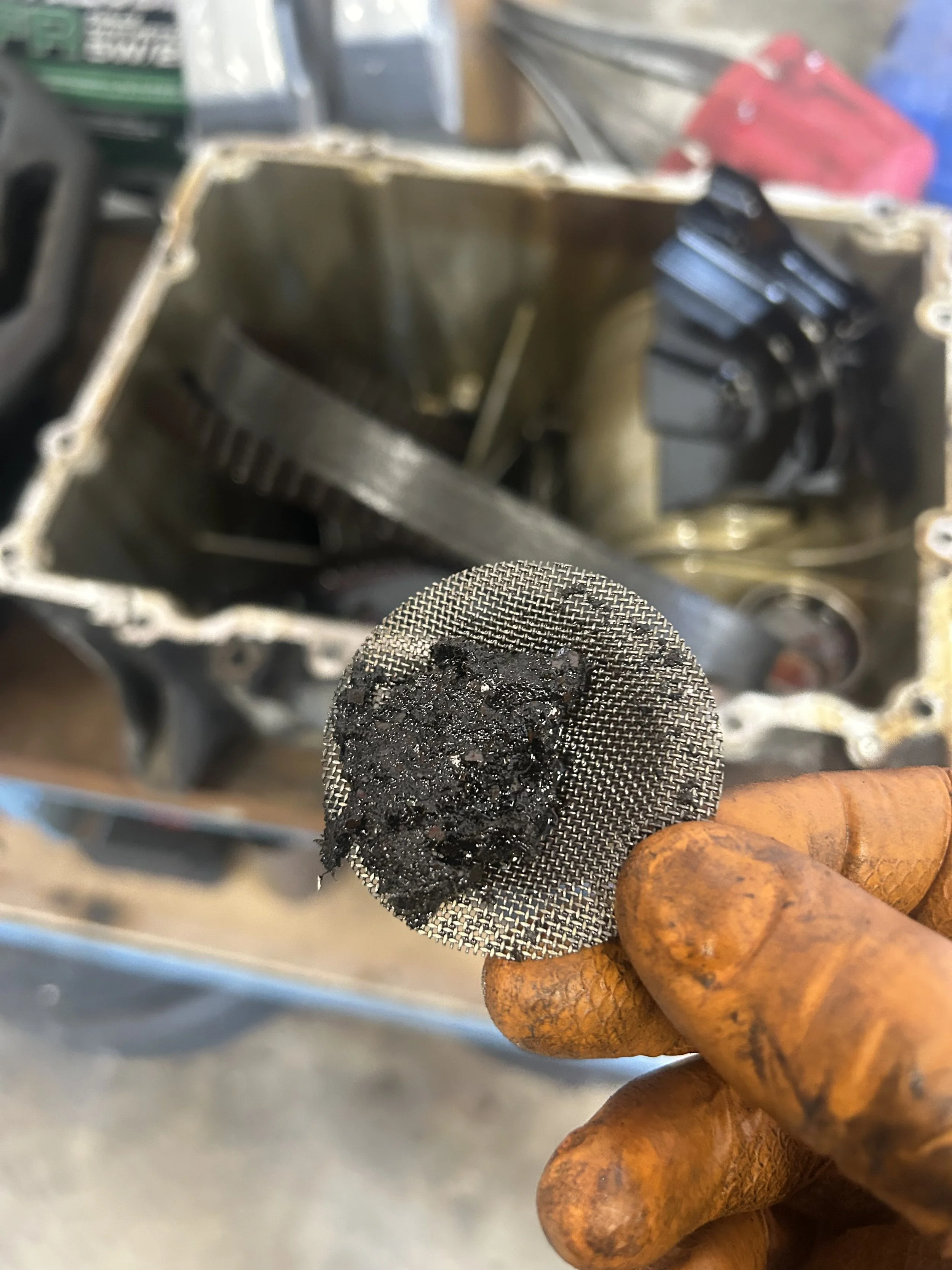 A gloved hand holding a small mesh screen with burnt debris on it, with an open engine component in the background.
