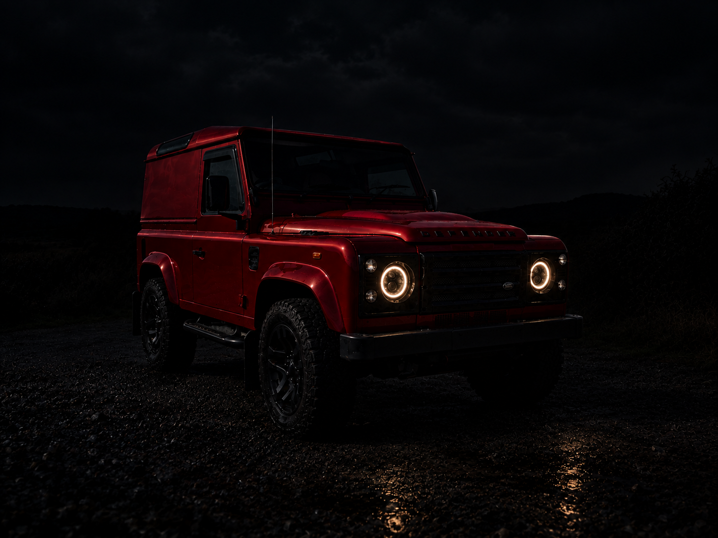 Red off-road vehicle at night with illuminated circular headlights, dark sky with clouds, and rugged terrain.