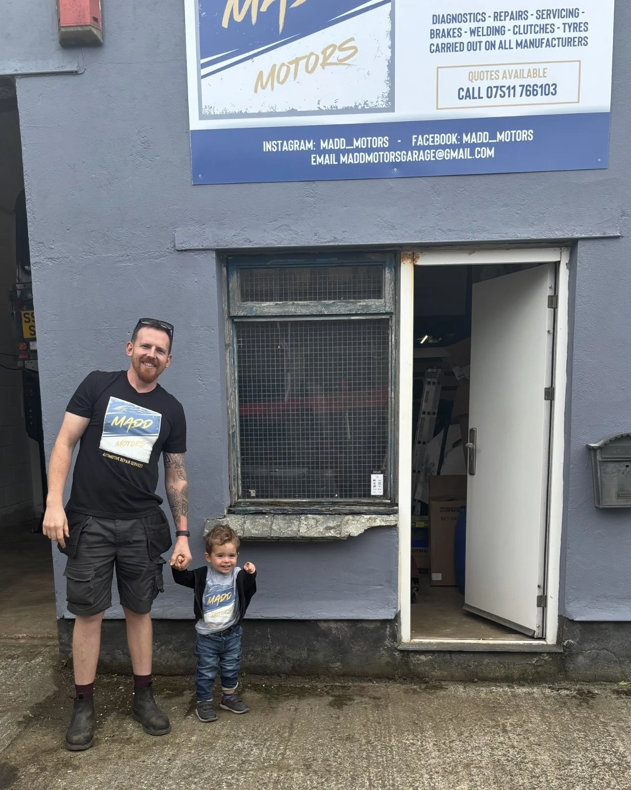 A man and a young boy standing outside a garage with a gray exterior wall, a window, and an open door. The man has a beard, short hair, and tattoos on his arm, wearing a black Madd Motors T-shirt, shorts, socks, and boots. The boy is smiling, has short hair, and is wearing a Madd Motors T-shirt and jeans. There is a large sign above the garage advertising Madd Motors' automotive repair services, with contact information and social media handles.
